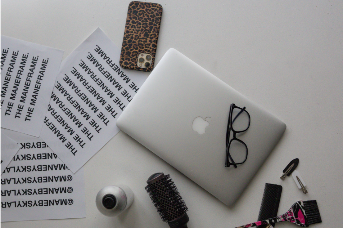 Clothing and electronic accessories on a white surface, including a closed silver MacBook with glasses on top, a leopard print phone case, a laptop cleaning brush, a small bottle of cleaning spray, black hair clips, adhesive labels with 'THE MANEFRAME' printed on them, and a pink and black hairbrush.