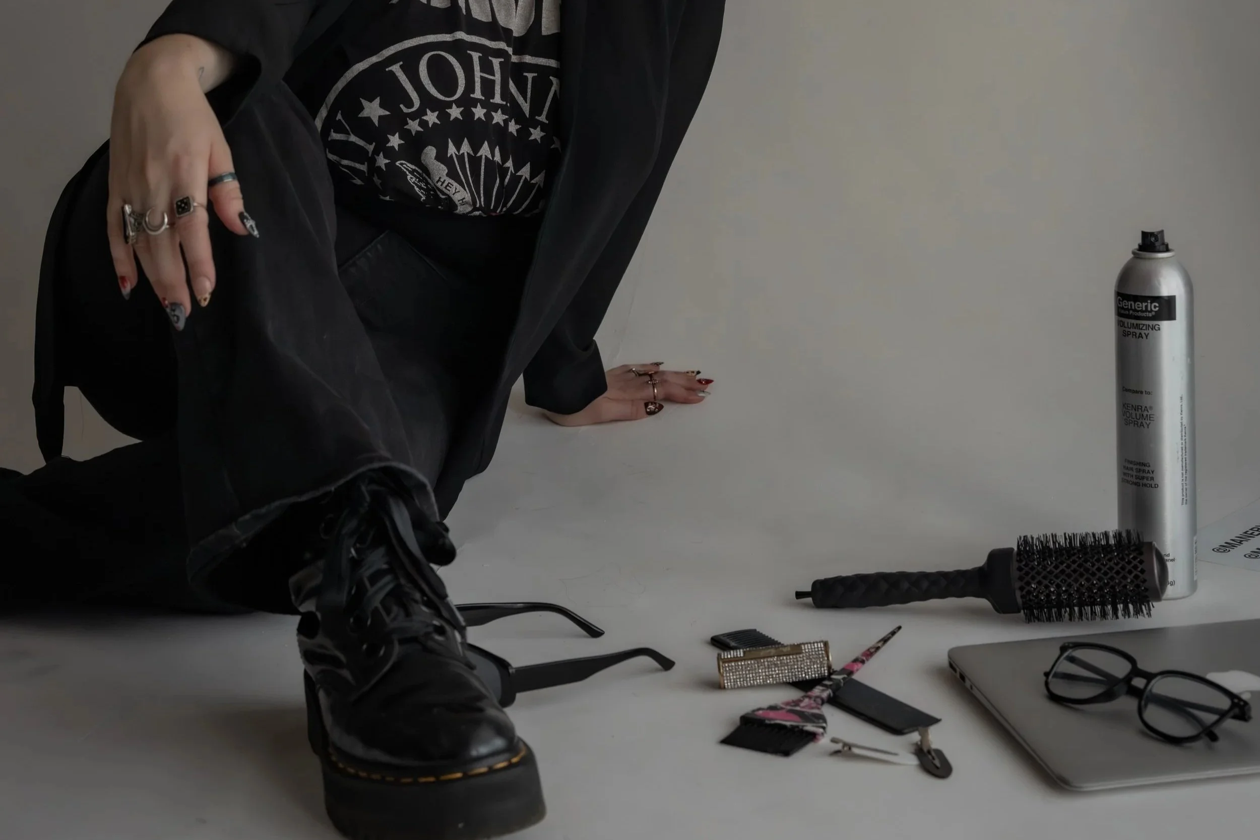 Person in black clothing kneeling with a hand on the ground, surrounded by hair styling tools including a round brush, hairspray, and a hair clip, on a white surface.
