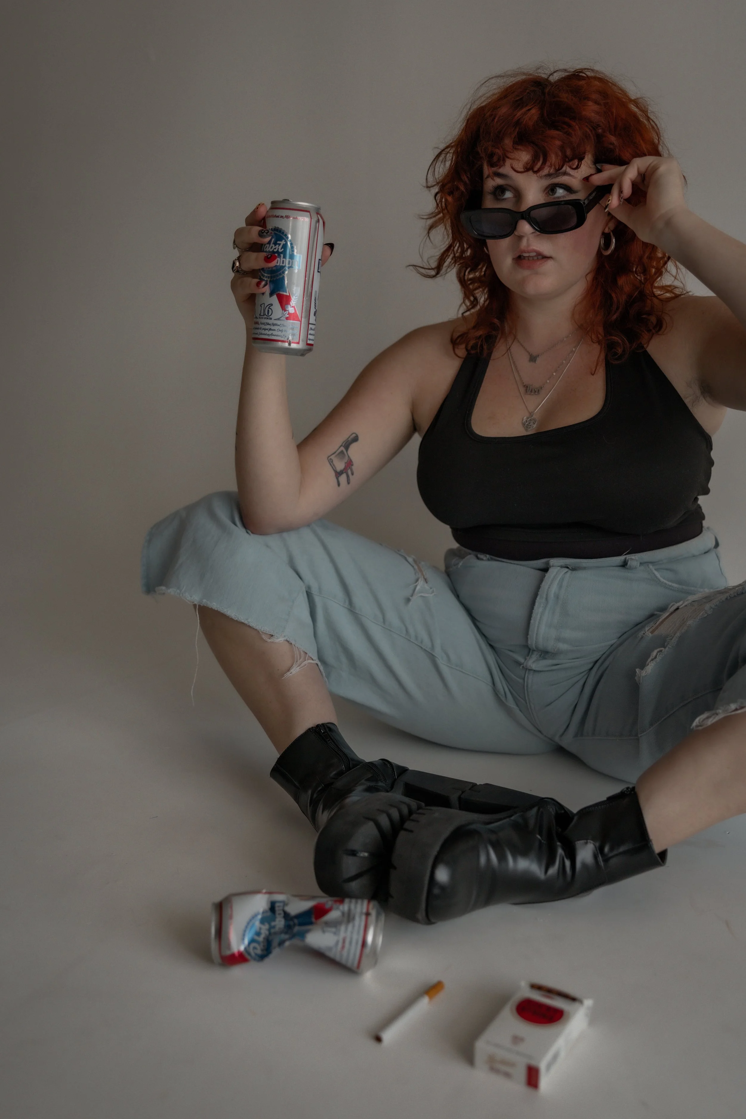 A woman with curly red hair sitting on the floor, holding a beer can, wearing sunglasses, a black tank top, ripped jeans, and black boots, with a cigarette and a pack on the ground.