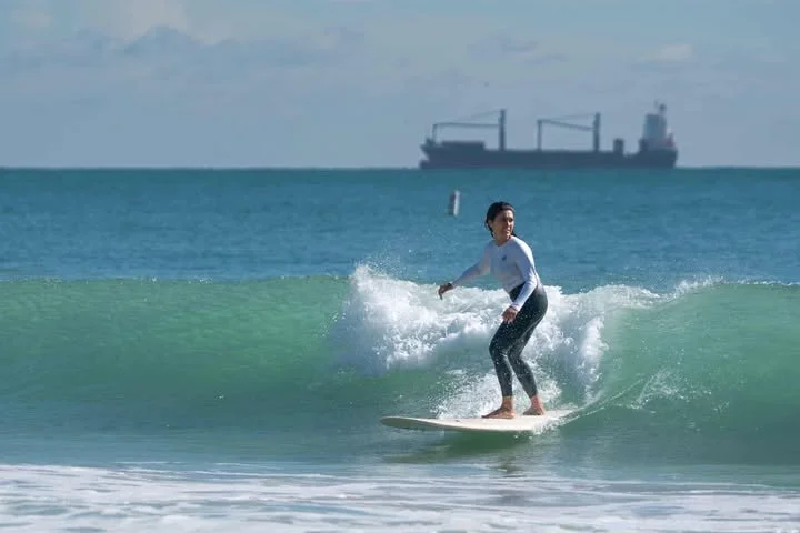 A woman surfing on a small wave near the shore with a large cargo ship in the background during daytime.