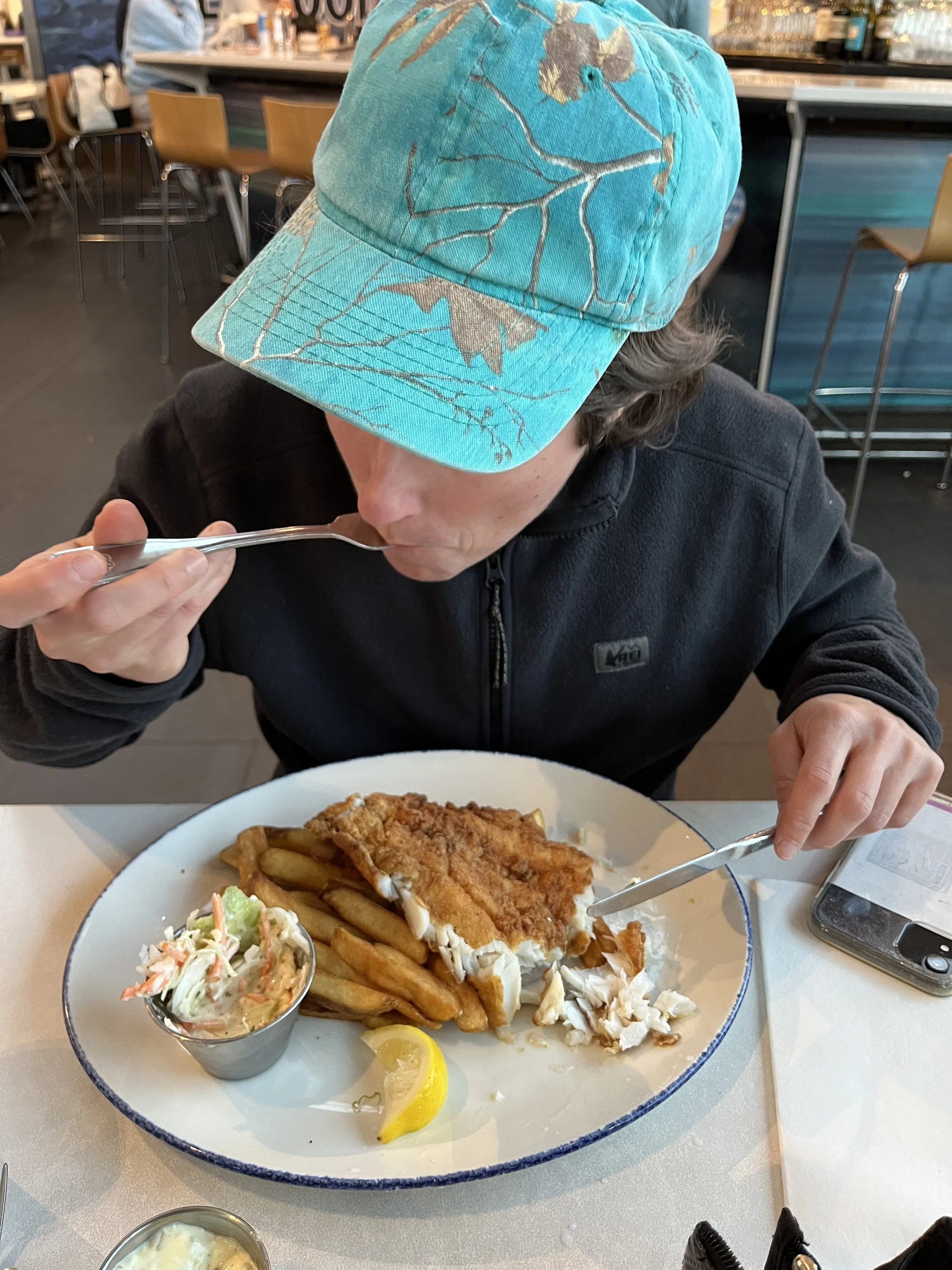 A person wearing a blue cap with leaf patterns is eating fish and chips at a restaurant.