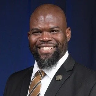 A smiling man with a beard, wearing a suit and tie, against a blue background.