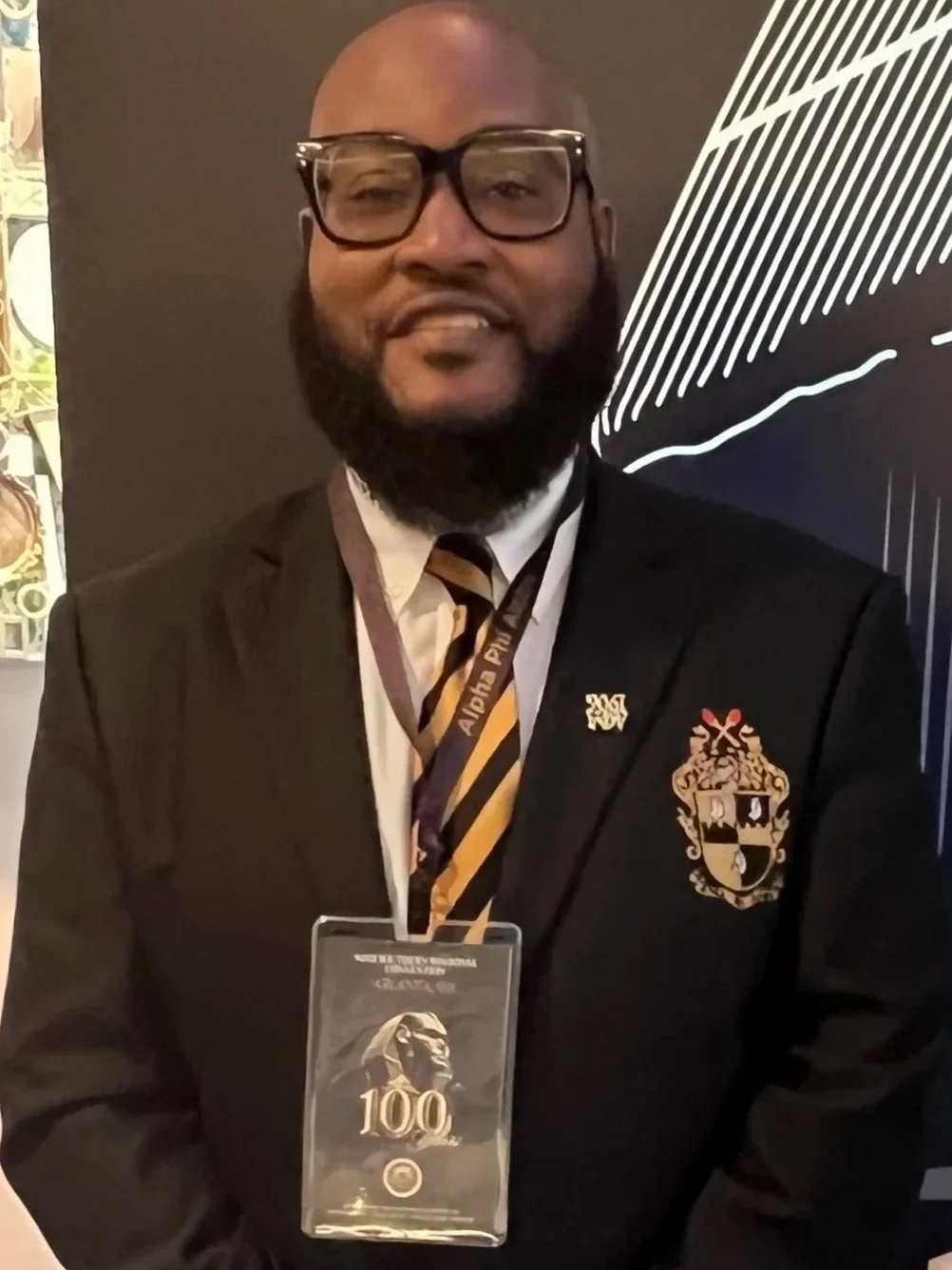A man with glasses and a beard wearing a black suit, white shirt, and a yellow and black striped tie, stands in front of a black background with white line art. He has a badge hanging around his neck with the number 100 and a crest on his lapel.