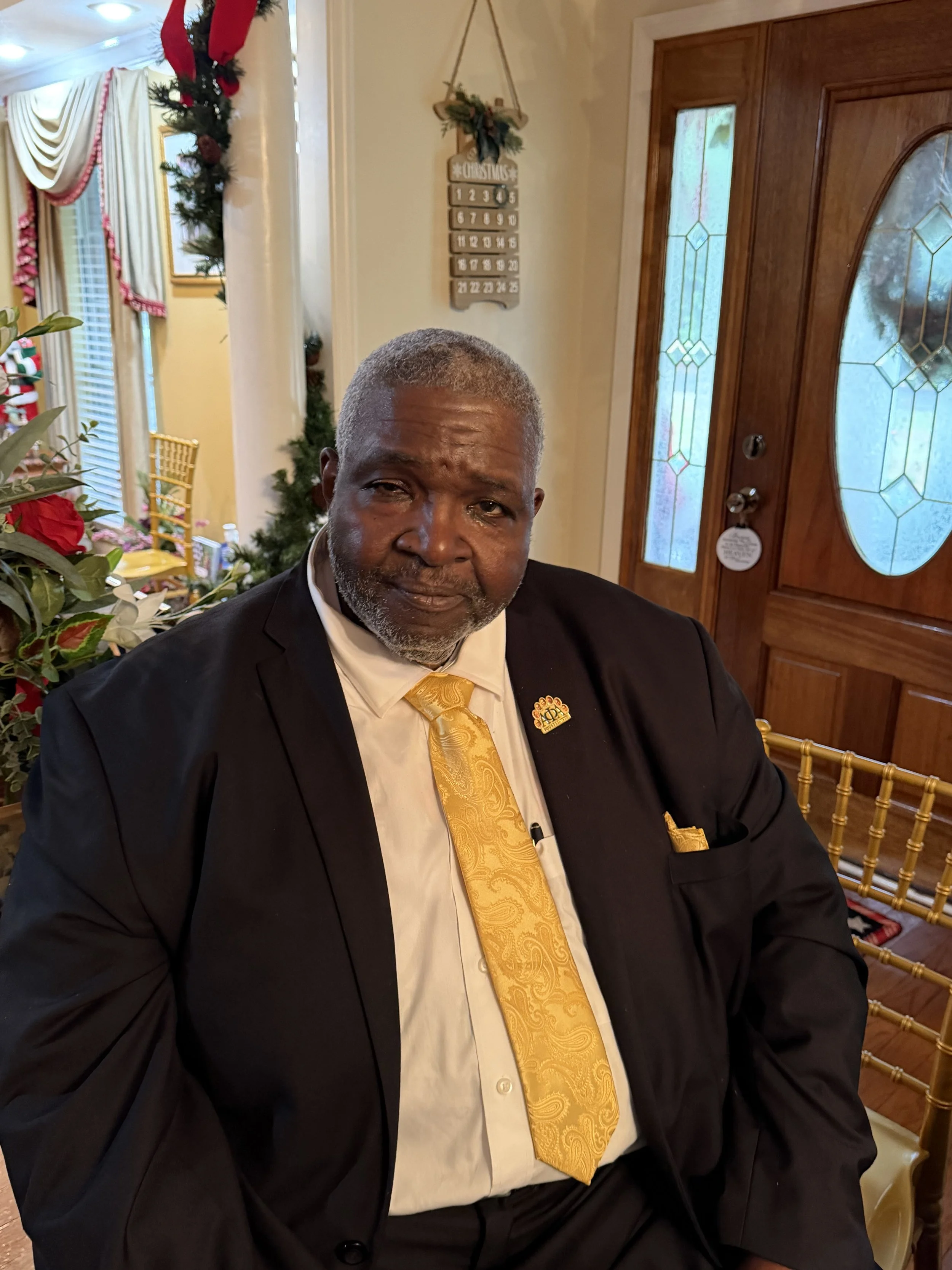 A mature man with gray hair and beard dressed in a black suit, white shirt, and gold tie, seated indoors during Christmas time, with Christmas decorations and a decorated door in the background.
