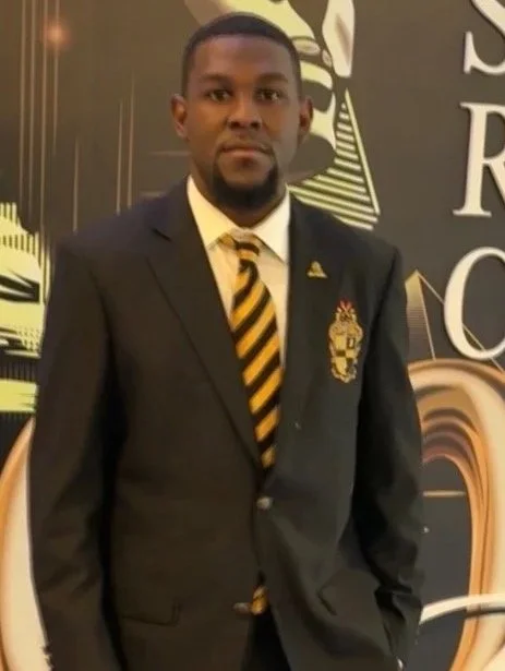 A man in a black suit with a yellow and black striped tie, standing indoors against a decorative backdrop.