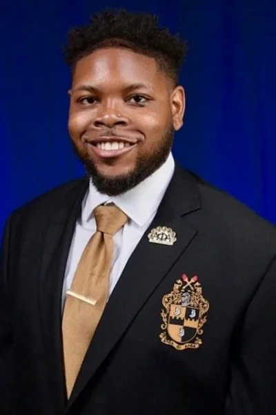 A professional man with short curly hair and a beard, smiling and wearing a dark suit, white shirt, gold tie, and a crest emblem on his blazer, standing against a blue background.