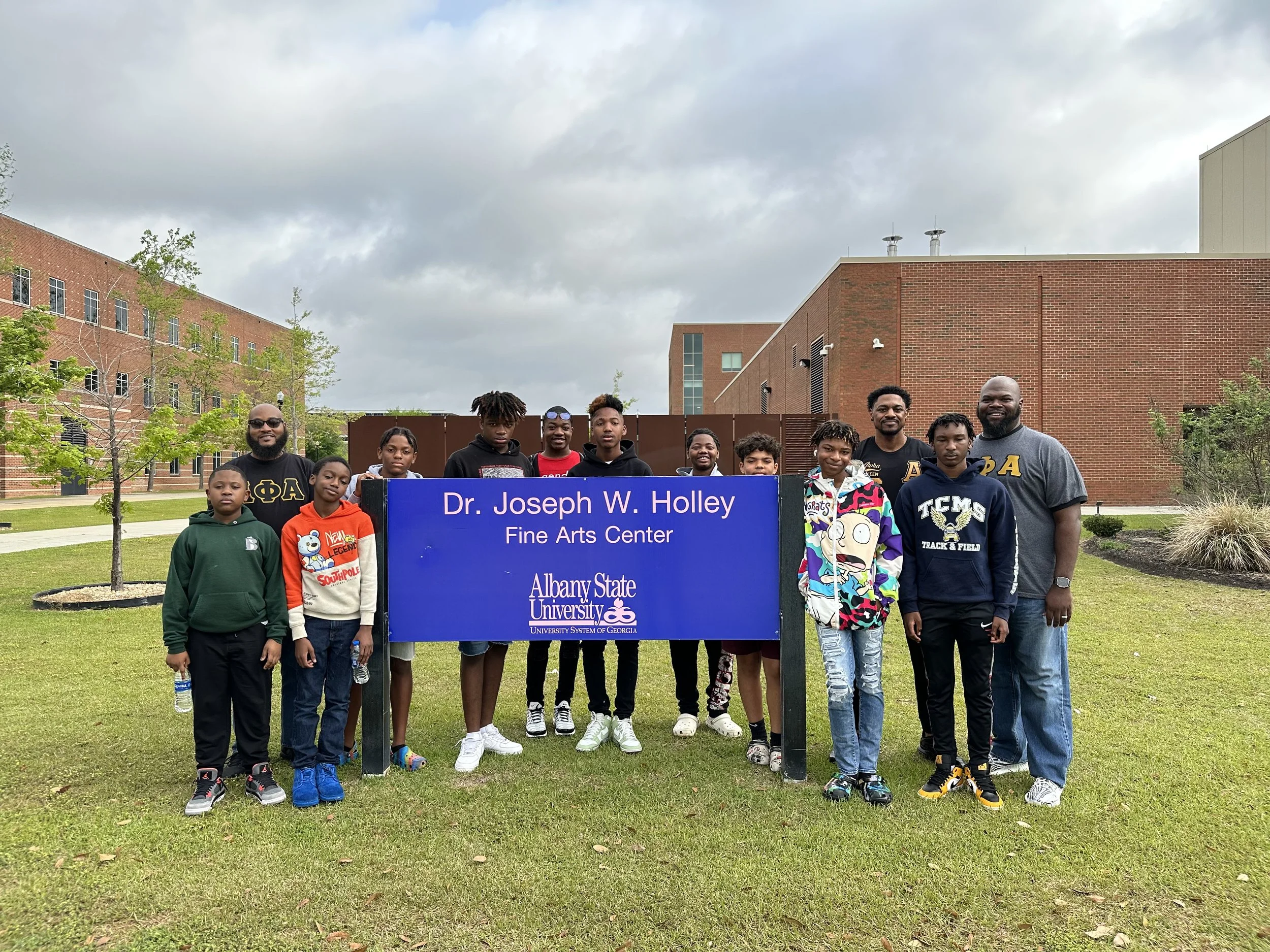 Group of children and adults standing outdoors in front of a blue sign for Dr. Joseph W. Holley Fine Arts Center at Albany State University, on a grassy area with trees and buildings under a cloudy sky.