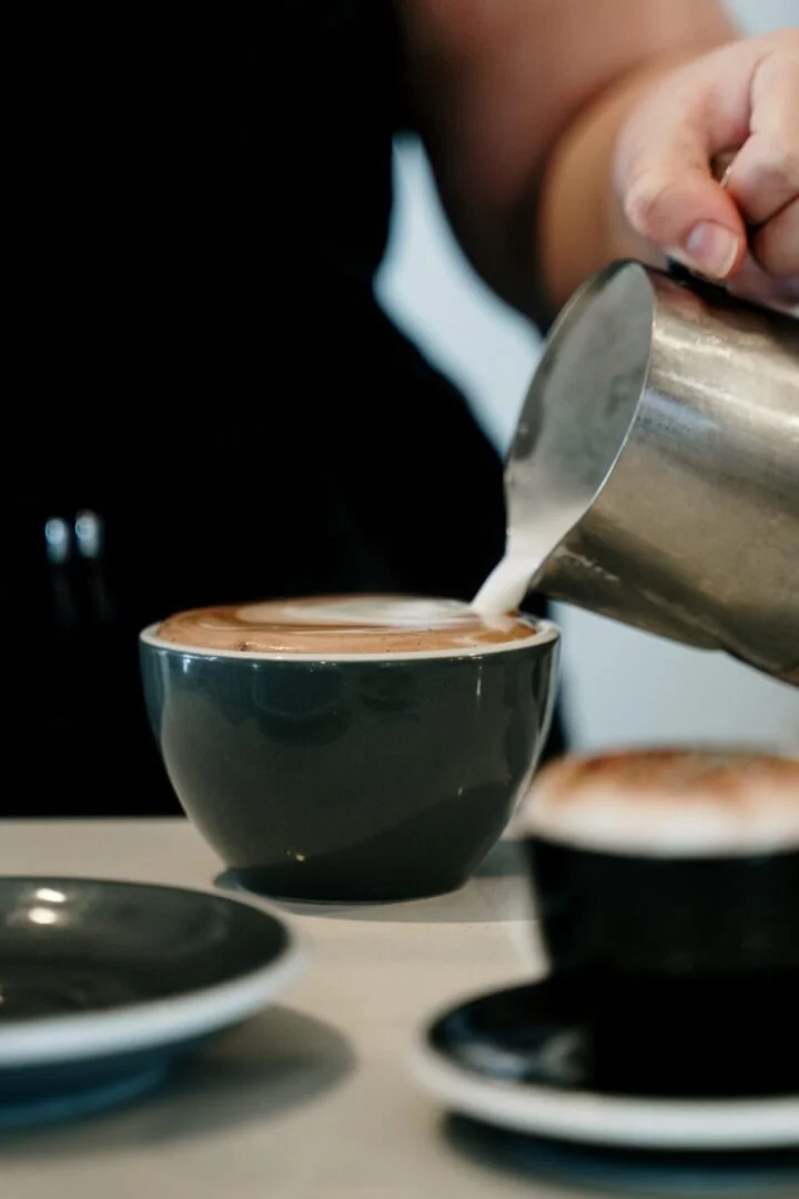 A person pouring steamed milk into a dark gray coffee cup to make a latte, with other cups and saucers on the table.