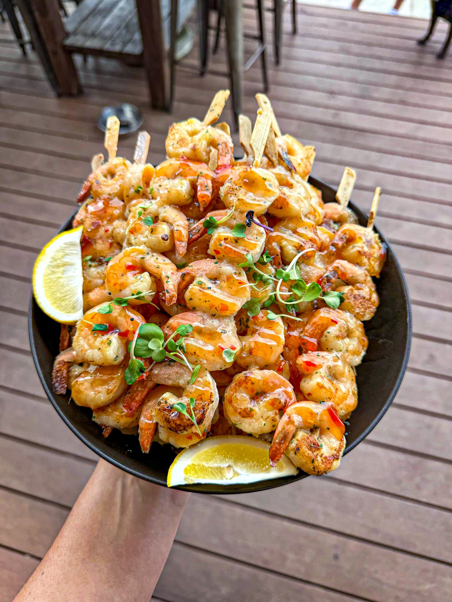 A black bowl filled with grilled shrimp garnished with microgreens, with lemon wedges on the side, held over a wooden deck patio.