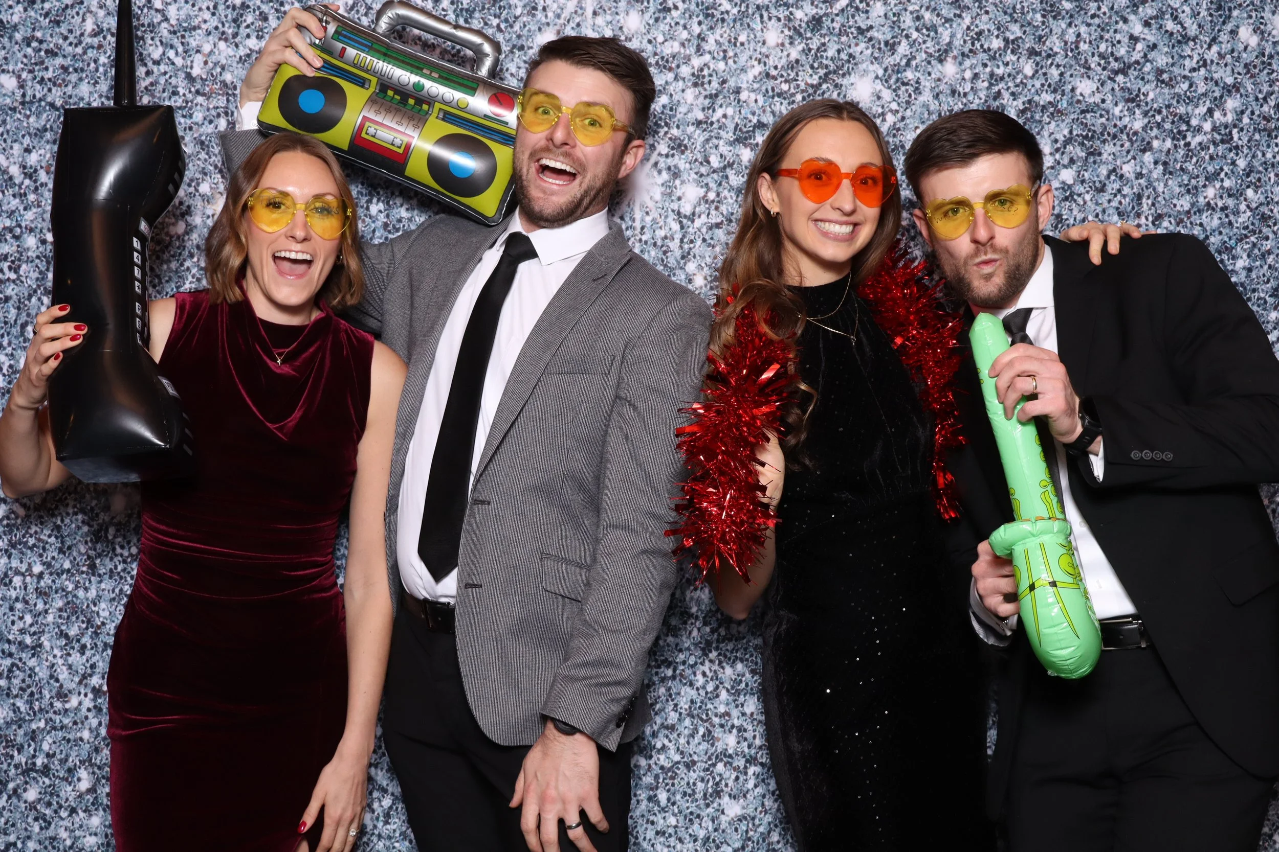 Group of four people at a party wearing colorful glasses and holding inflatable party props, with a speckled backdrop.