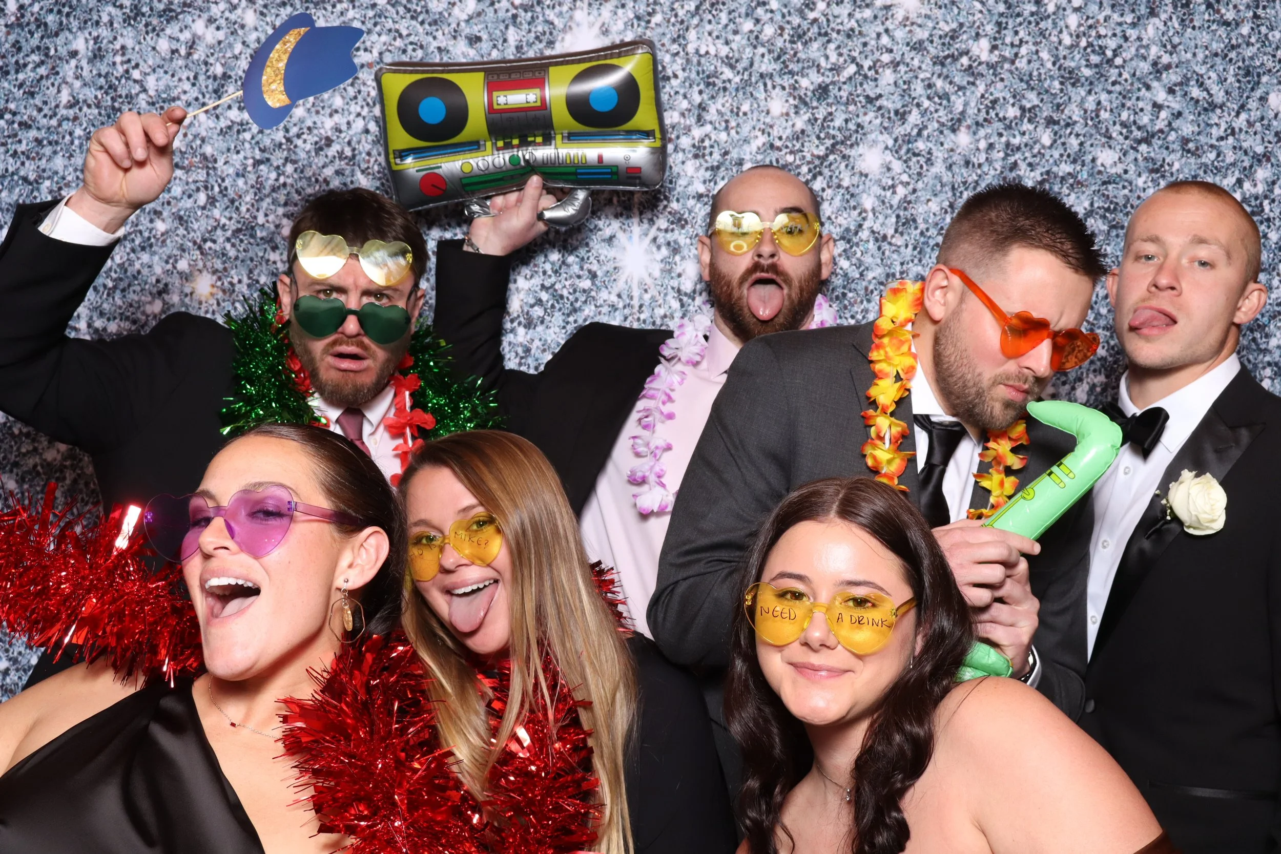 Group of people at a party wearing colorful accessories, sunglasses, and costumes, posing against a sparkly backdrop.