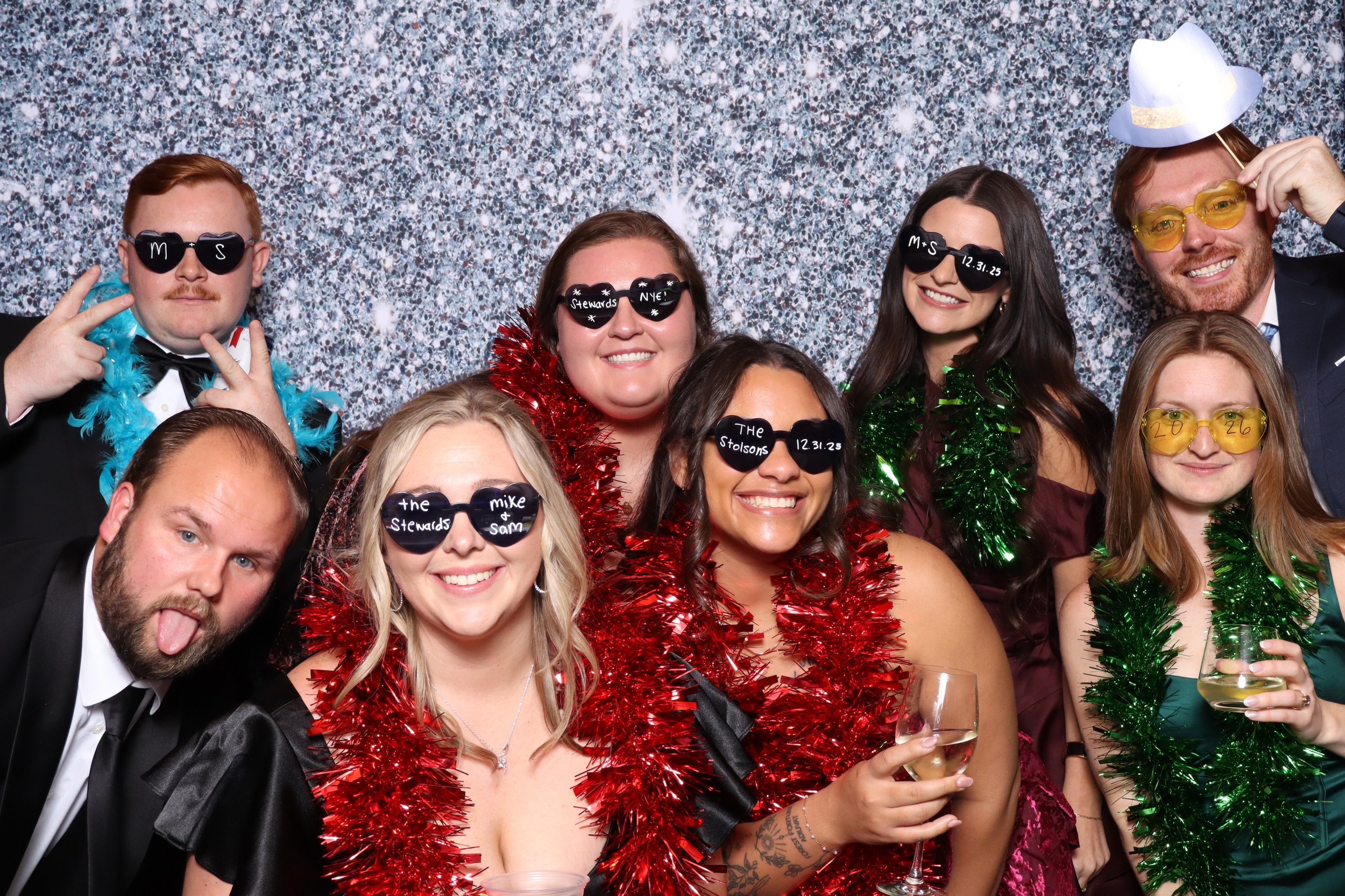 Group of eight people dressed in formal attire at a festive party, wearing novelty sunglasses and colorful tinsel boas, posing cheerfully in front of a sparkly silver backdrop.