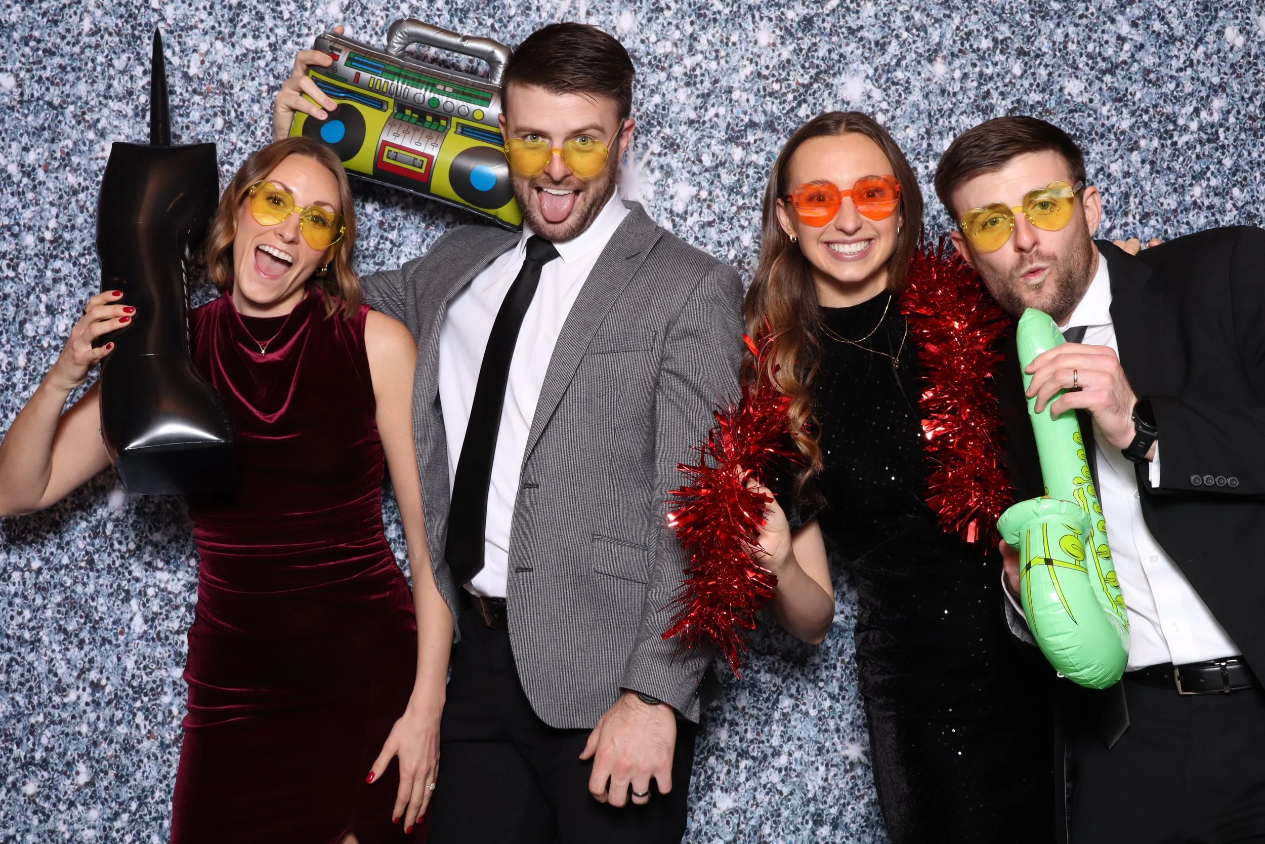 Four people at a party wearing colorful sunglasses, holding inflatable props, and posing with playful expressions against a glittery backdrop.