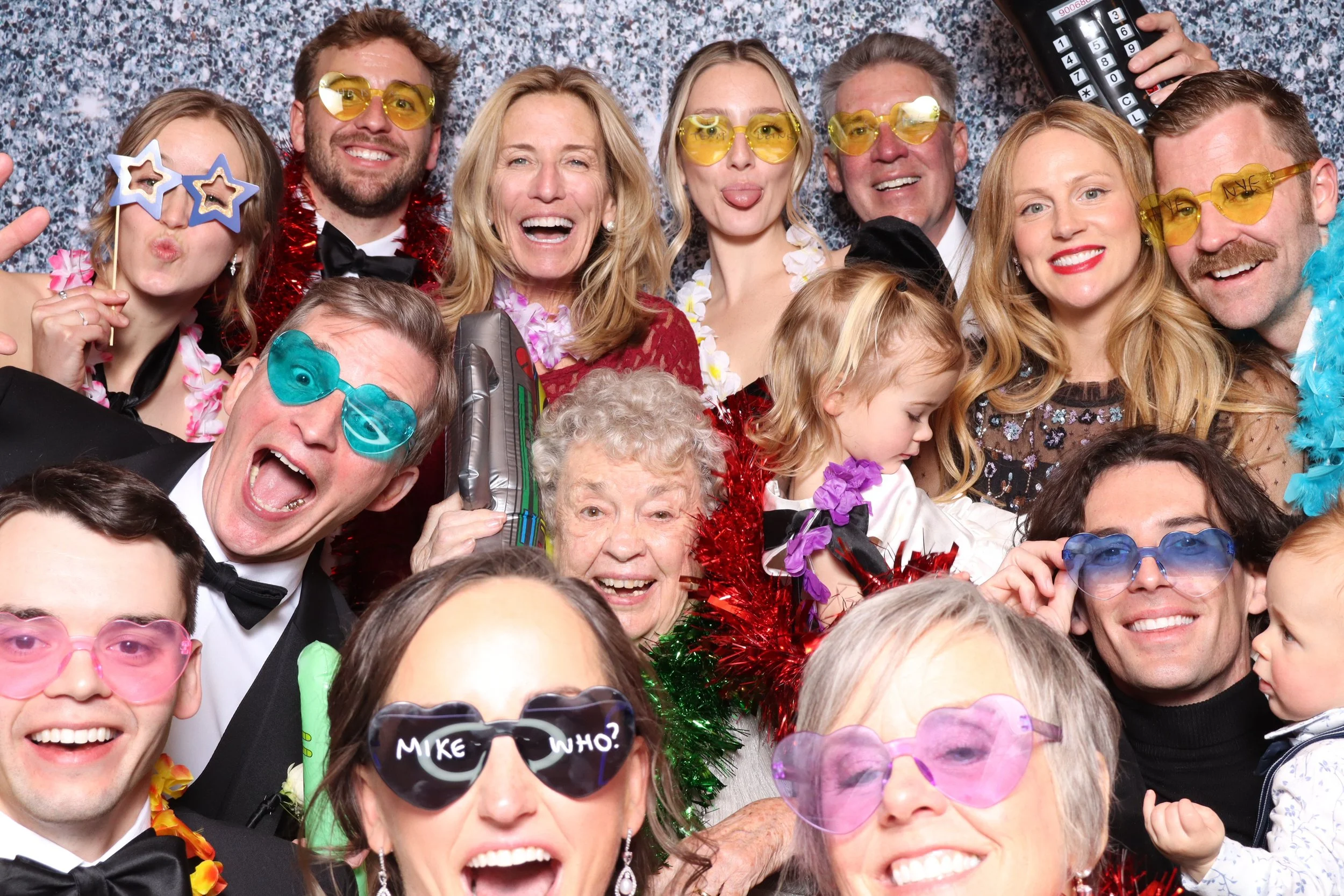 Group of people at a party wearing colorful sunglasses and costumes, celebrating with a festive background, some making funny faces, and holding party props.