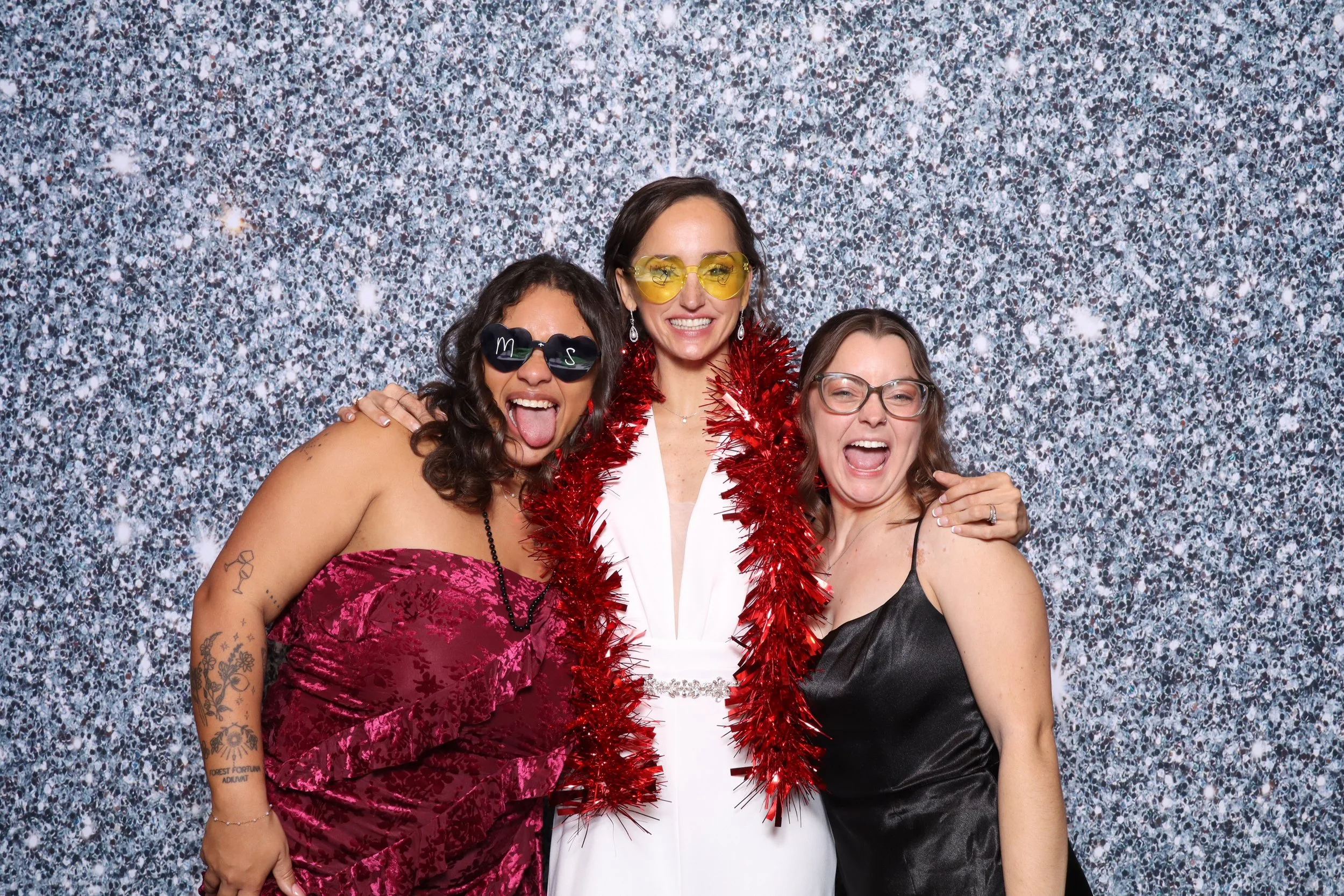 Three women celebrating at a party with a glittery silver background, wearing festive accessories and smiling.