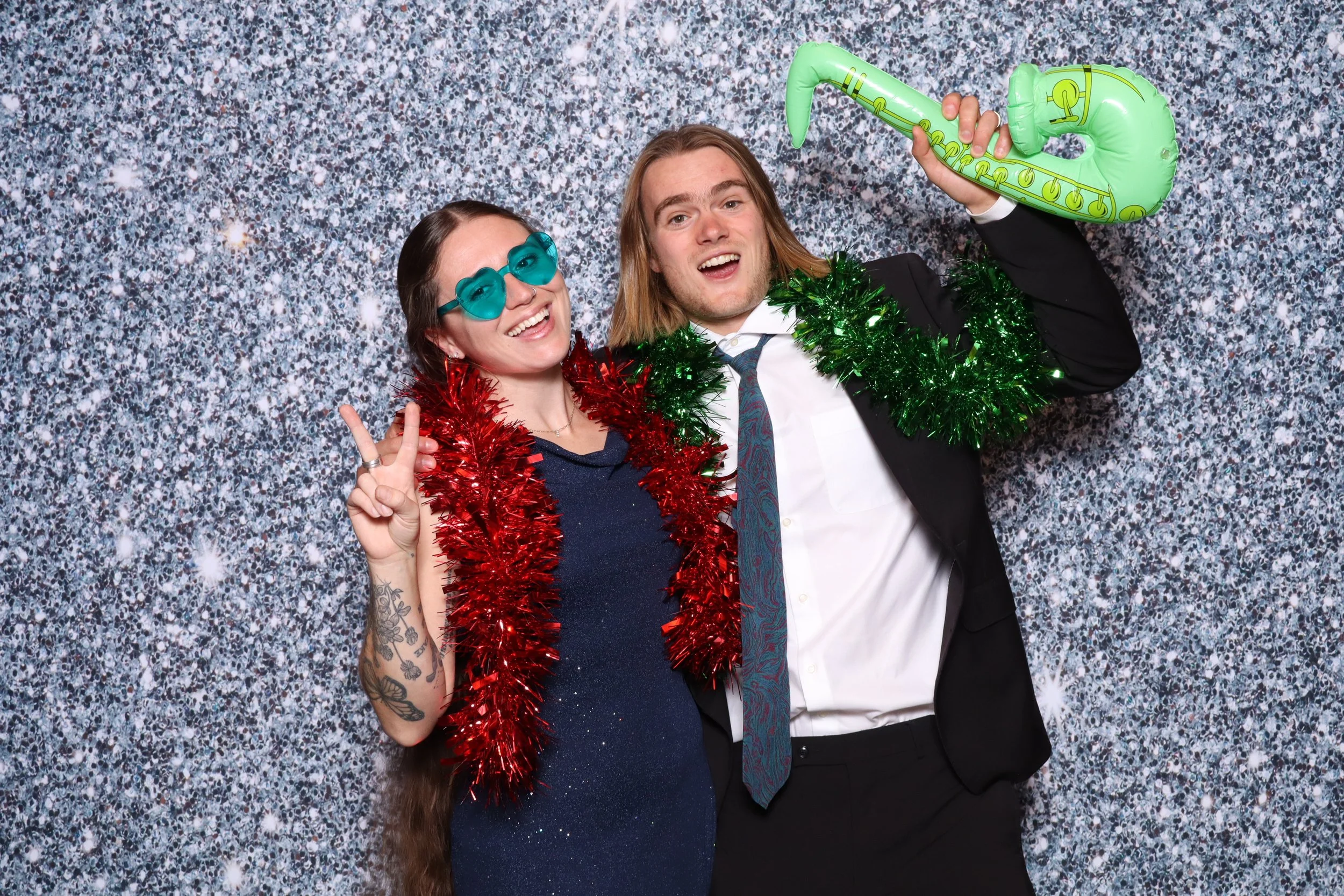 Two people celebrating in front of a sparkling silver backdrop, wearing holiday accessories including tinsel and sunglasses, with one holding a green inflatable saxophone.