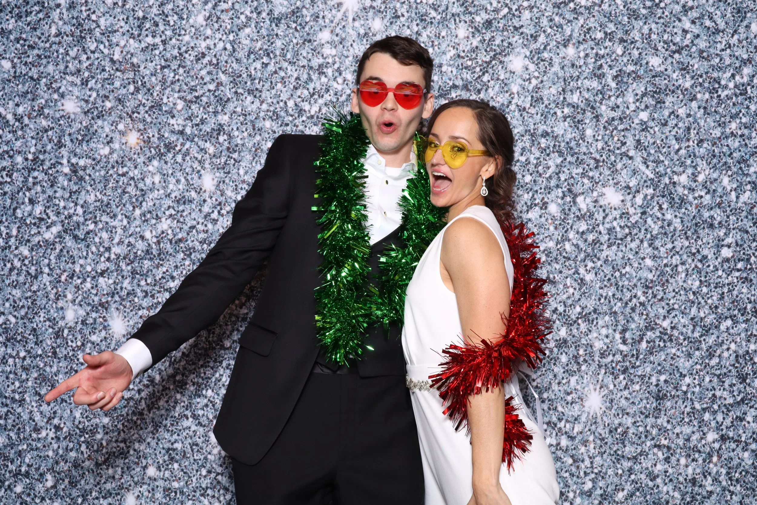 A man and woman dressed in festive attire at a party, wearing colorful sunglasses, with the man in a black suit and the woman in a white dress, both sporting tinsel boas against a glittery silver backdrop.
