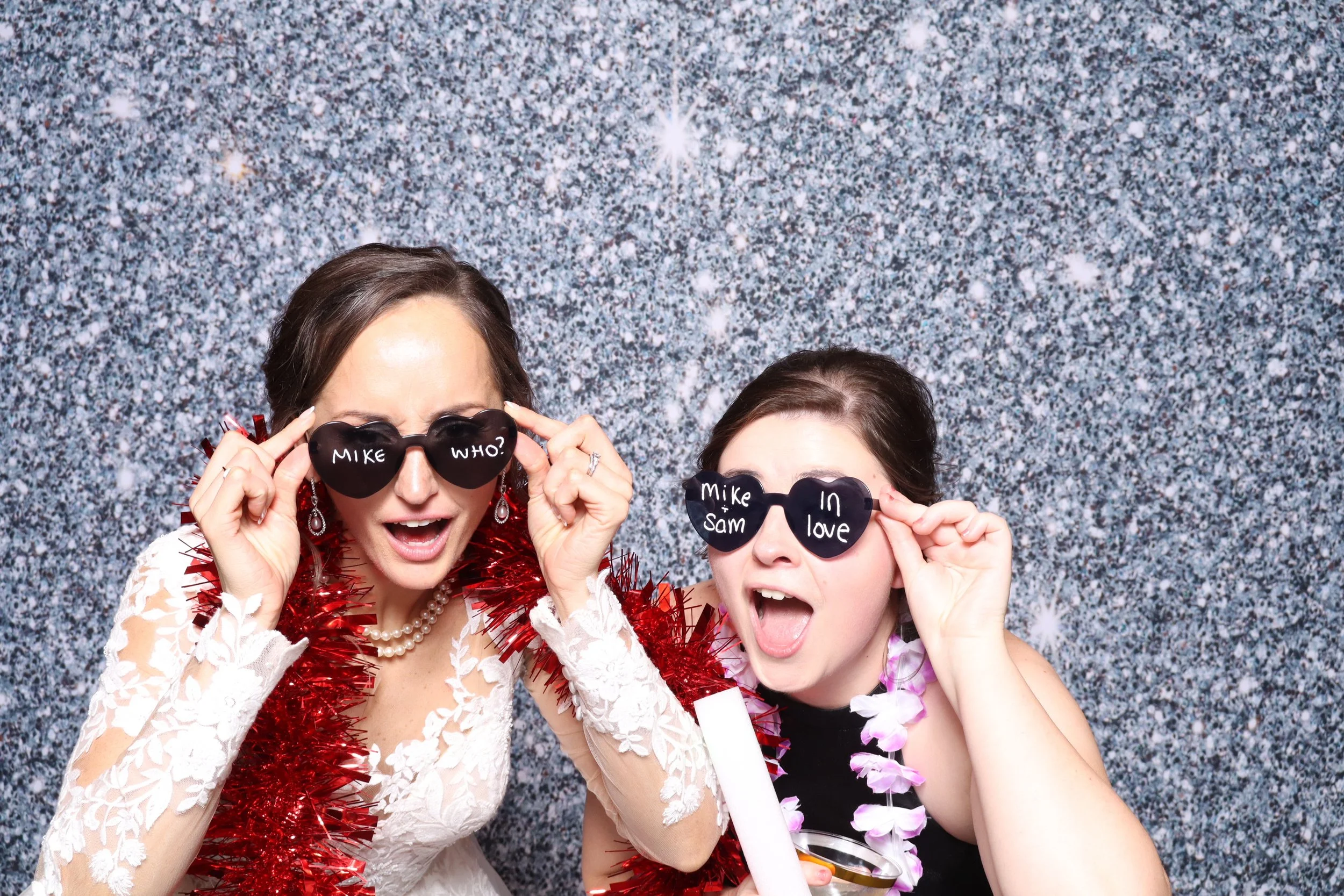 Two women dressed for a celebration, wearing heart-shaped sunglasses with messages written on them, and surrounded by festive accessories, standing in front of a sparkly silver background.