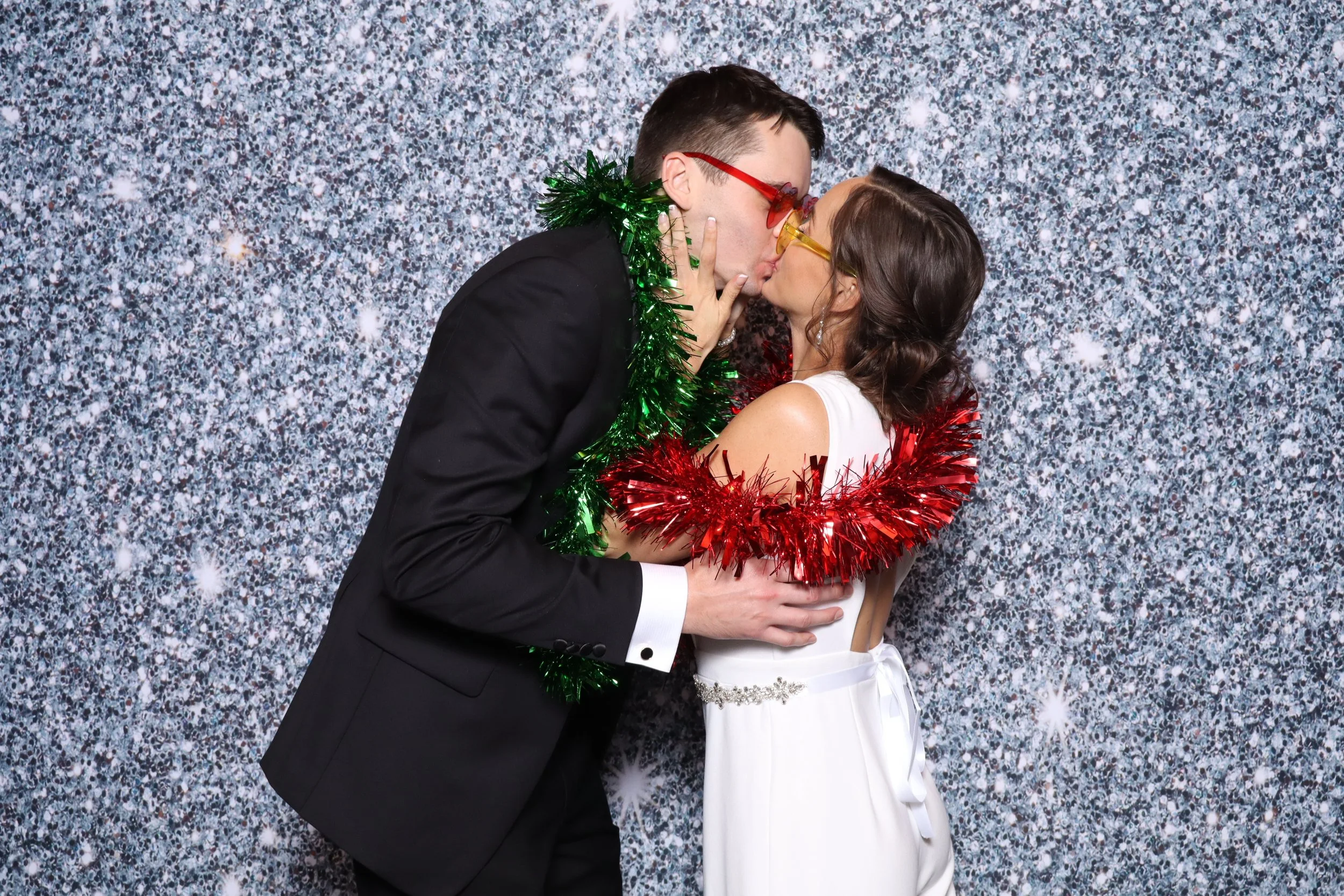 A man and woman kissing at a festive celebration, adorned with Christmas tinsel in green and red, in front of a silver glittery backdrop.