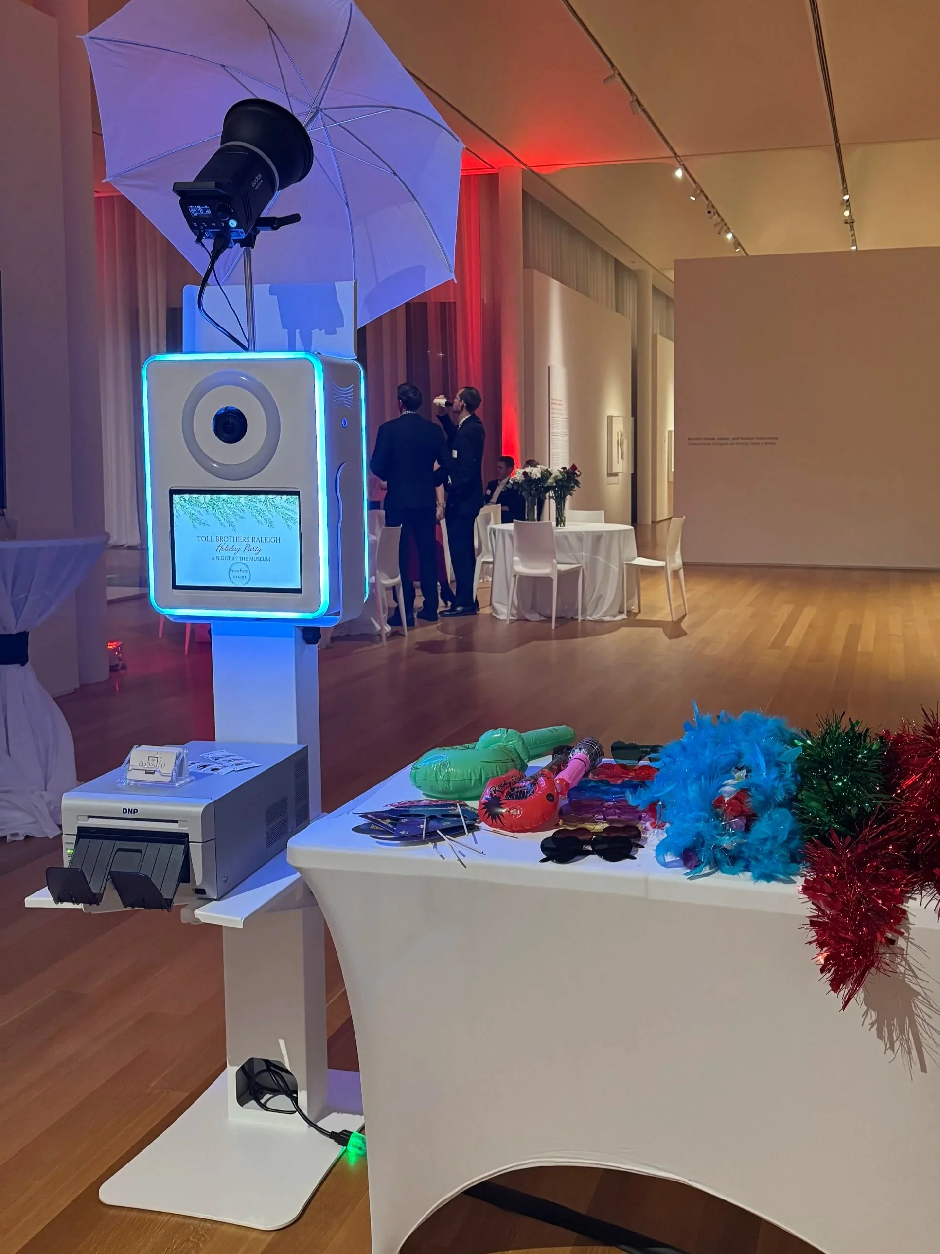 Photo of a photo booth setup with a large umbrella light, a camera on a stand, a mirror with a ring light, and a digital screen displaying a pink graphic for Cafécito Debelles Roastery in front of a staircase.