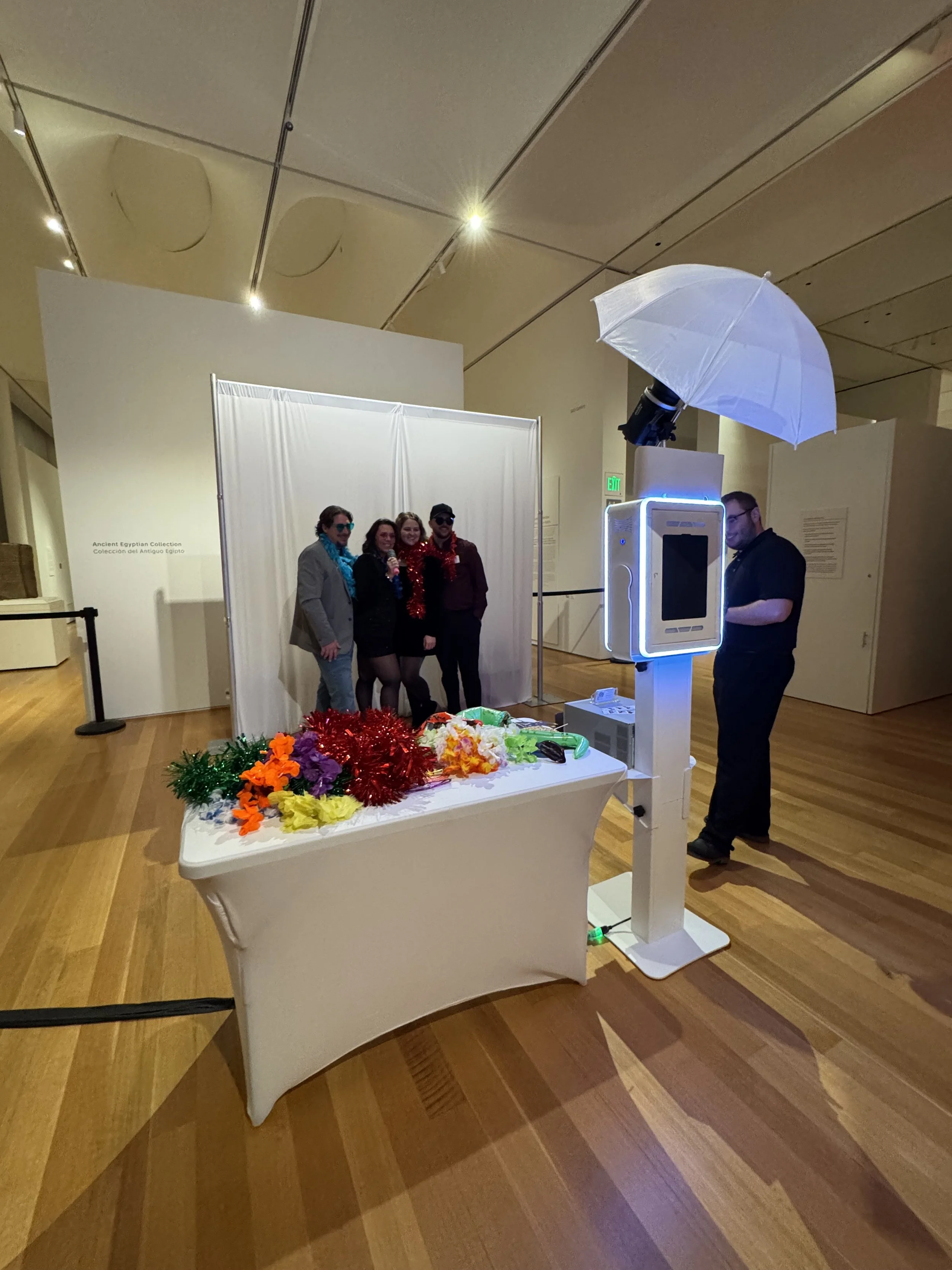 Photo of a photo booth setup with a large umbrella light, a camera on a stand, a mirror with a ring light, and a digital screen displaying a pink graphic for Cafécito Debelles Roastery in front of a staircase.