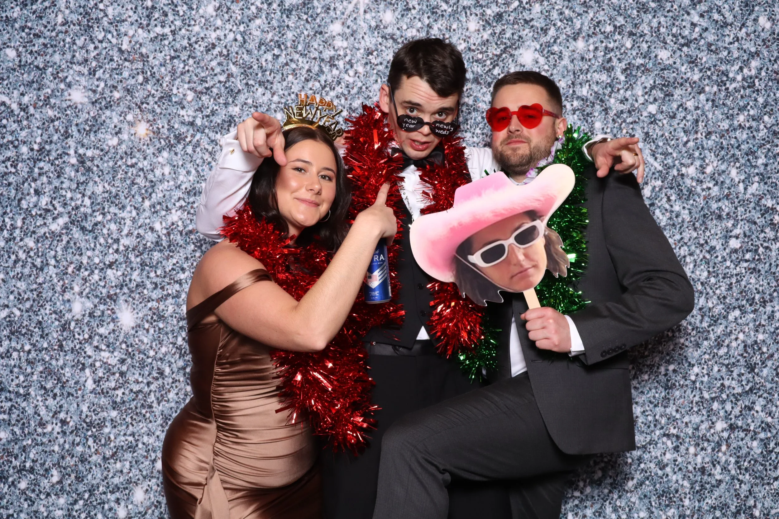 Four people at a party wearing fun costumes and accessories. The woman on the left is wearing a strapless satin dress with a 'Happy New Year' headband and a red tinsel scarf. The man in the middle is wearing oversized glasses with 'new year' written 