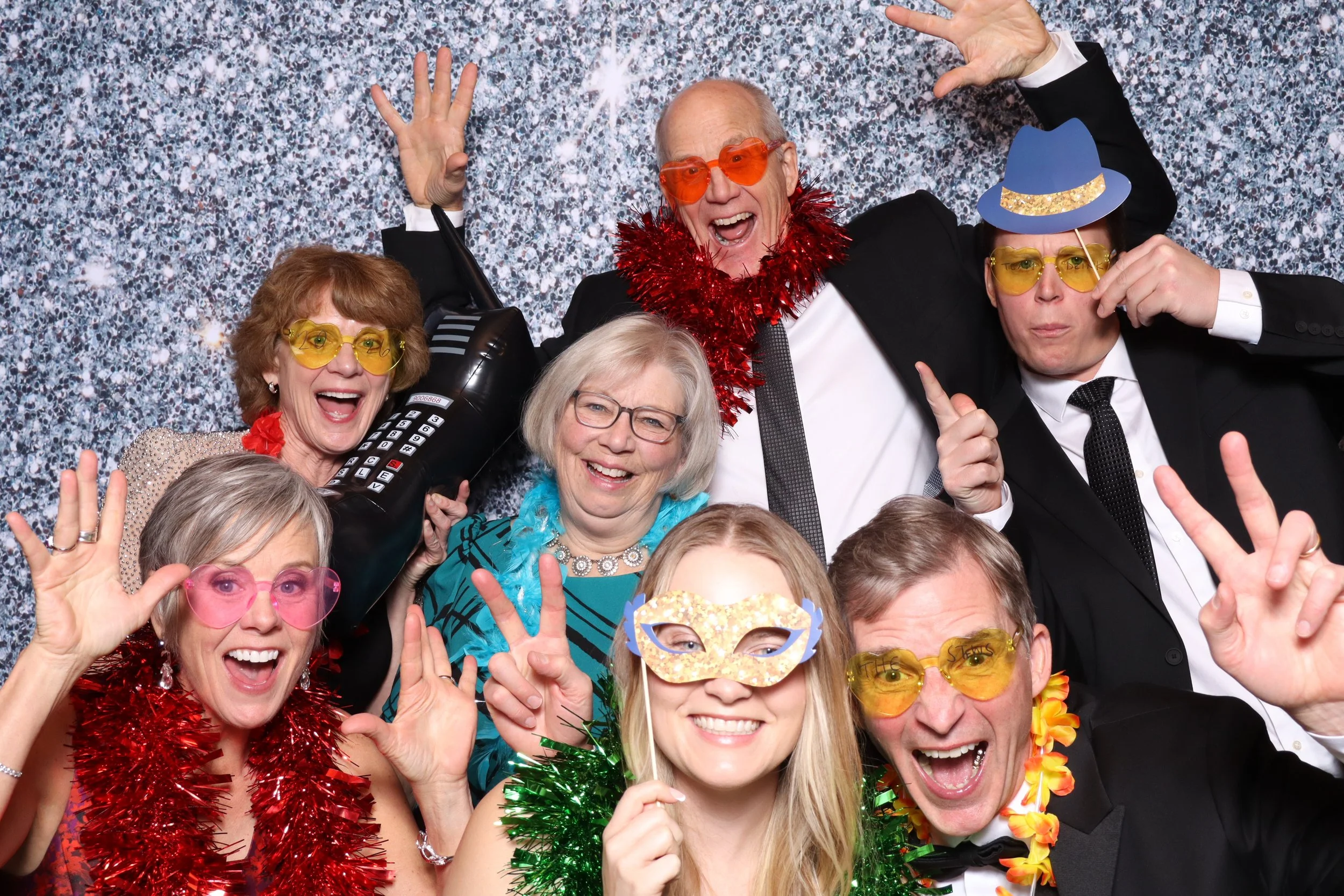 A group of cheerful people in party attire celebrating at a festive event, wearing colorful costumes, hats, glasses, and accessories, with a glittering backdrop.