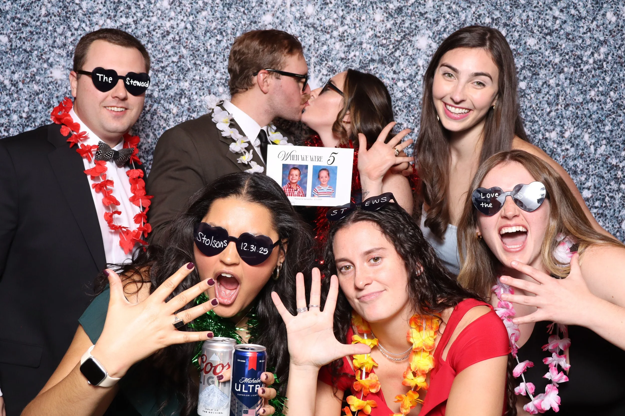 Group of people celebrating at a wedding, some wearing sunglasses with wedding-related writing, others wearing leis and holding drinks, in front of a silver glittery backdrop. One couple in the background is kissing, holding a sign that says 'When we