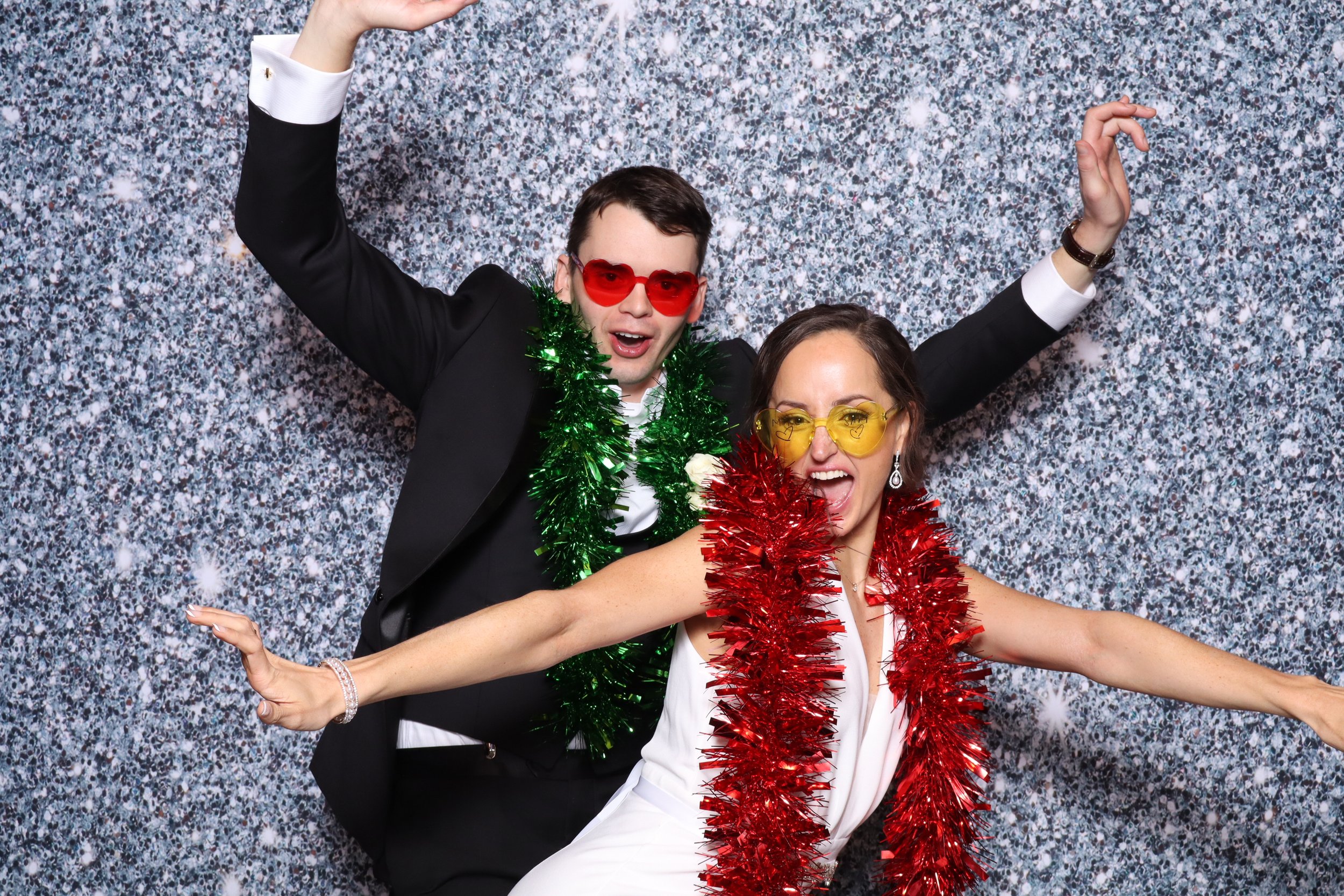Two people dressed in formal attire celebrating, wearing colorful holiday accessories like tinsel garlands and sunglasses, with a sparkling silver backdrop.