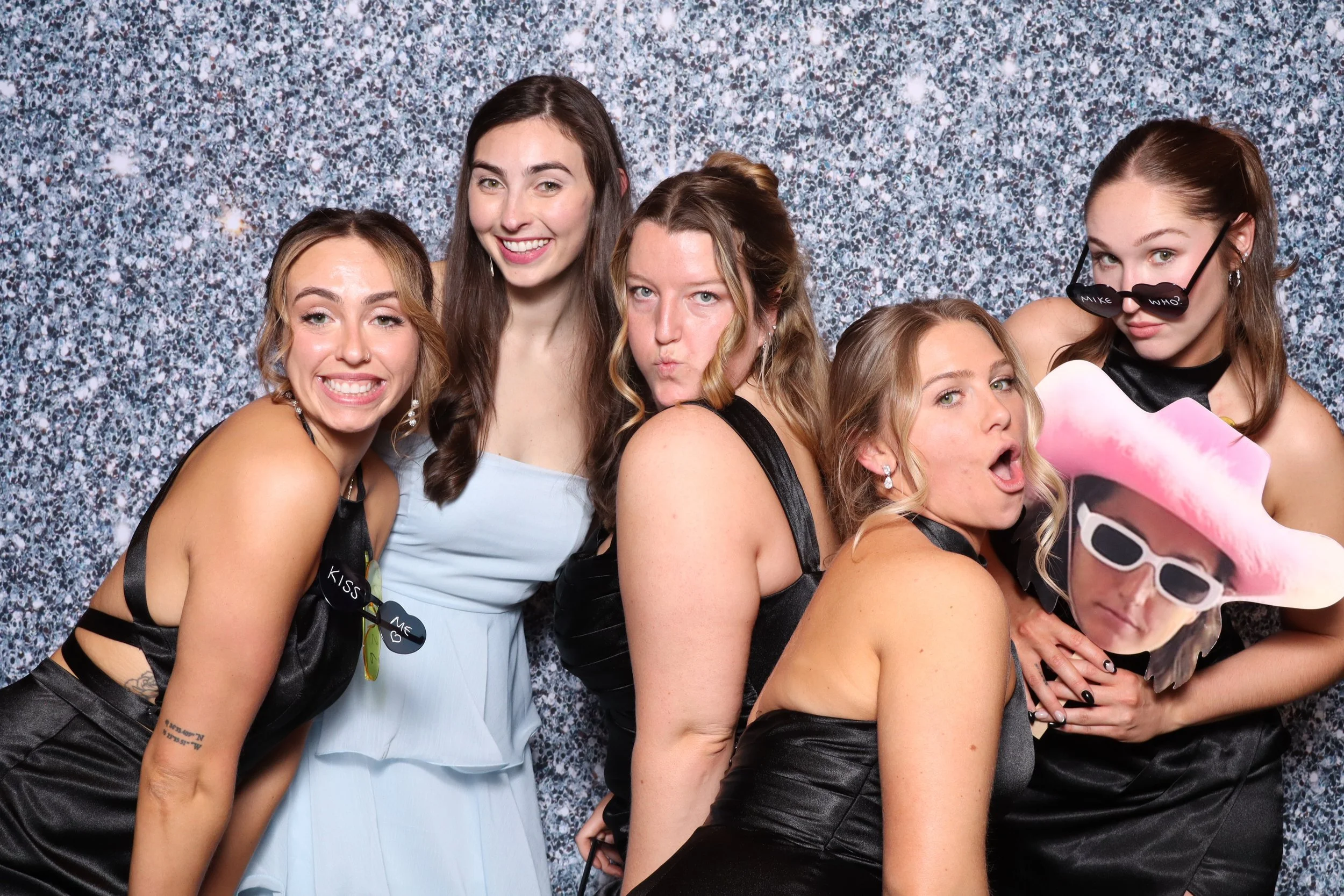 Six women at a party posing for a photo against a glittery background, wearing dresses and playful accessories like sunglasses and hats.