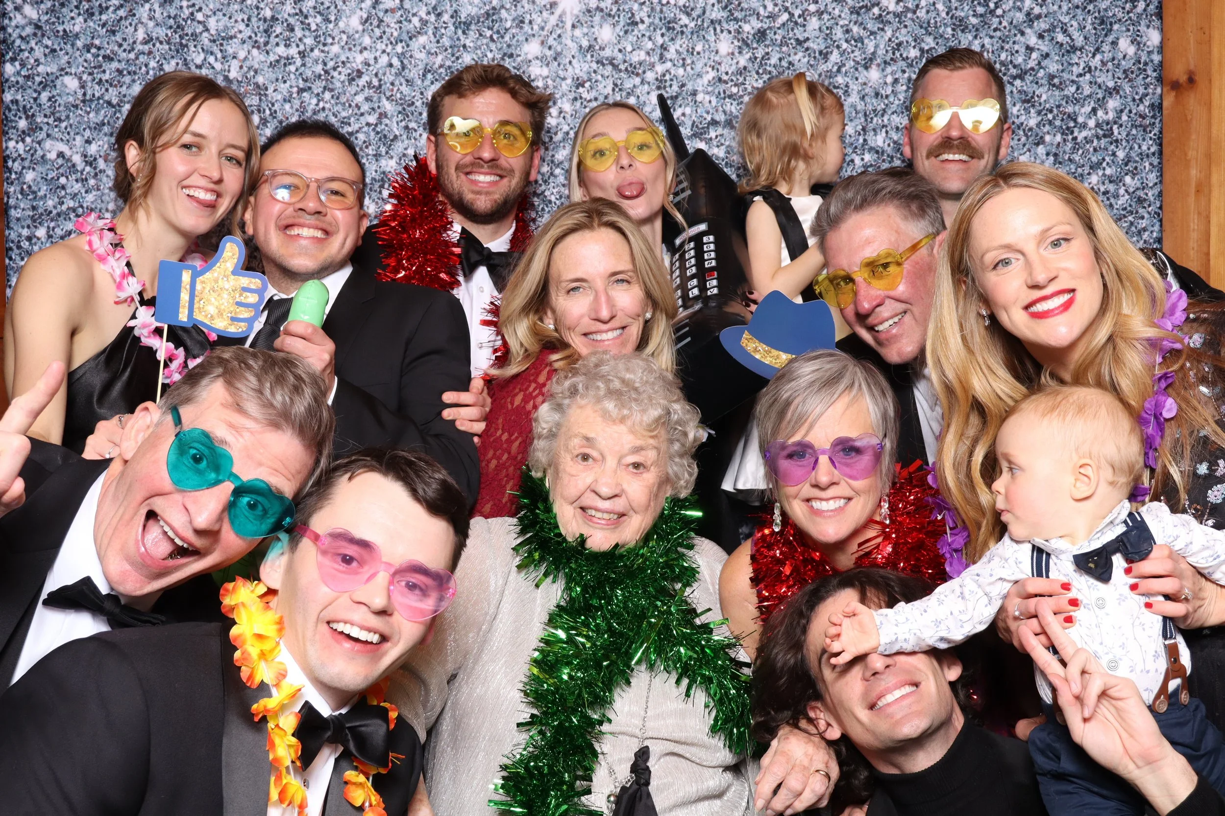 A group of people at a party, wearing colorful costume accessories, including sunglasses, leis, tinsel, and funny glasses, against a sparkling backdrop.