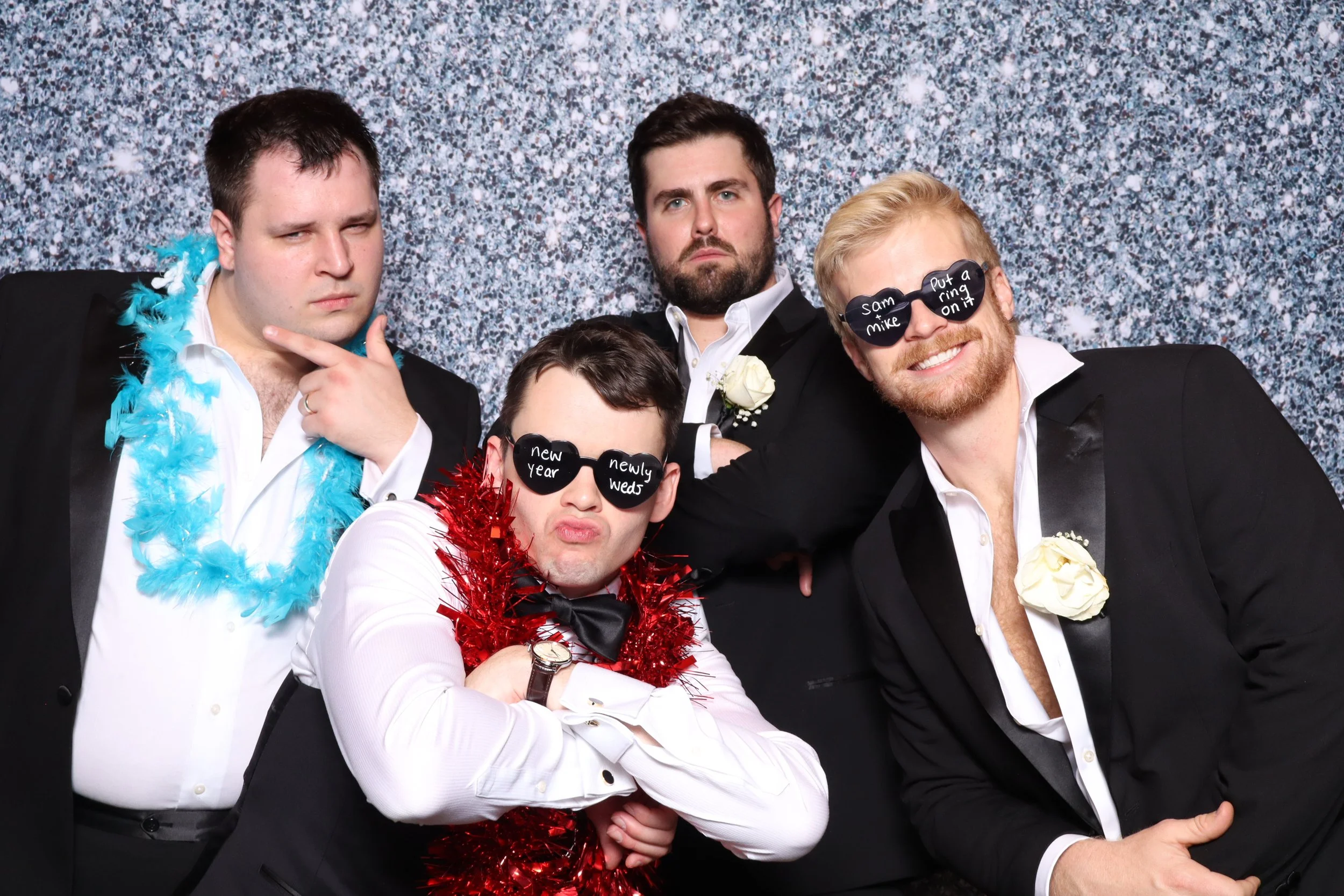 Four men in tuxedos posing with fun accessories in front of a glittery background, some wearing sunglasses with messages, some with boas and flowers.