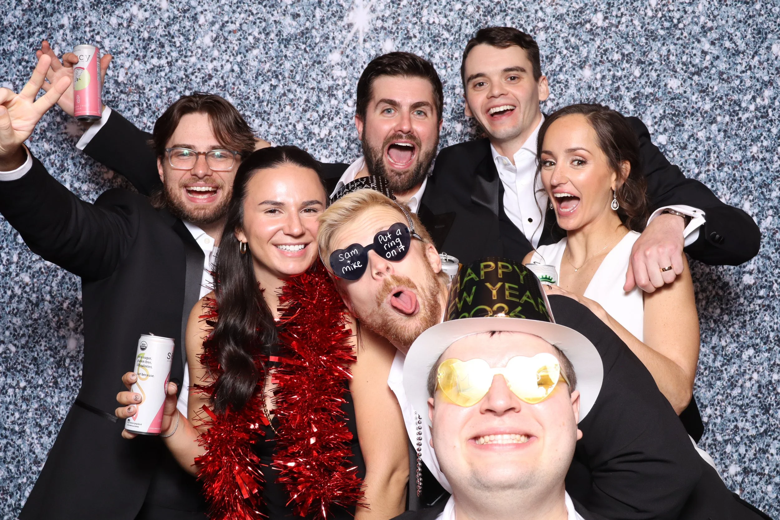 Group of seven people celebrating New Year's Eve, wearing party hats, glasses, and accessories, with a festive background.