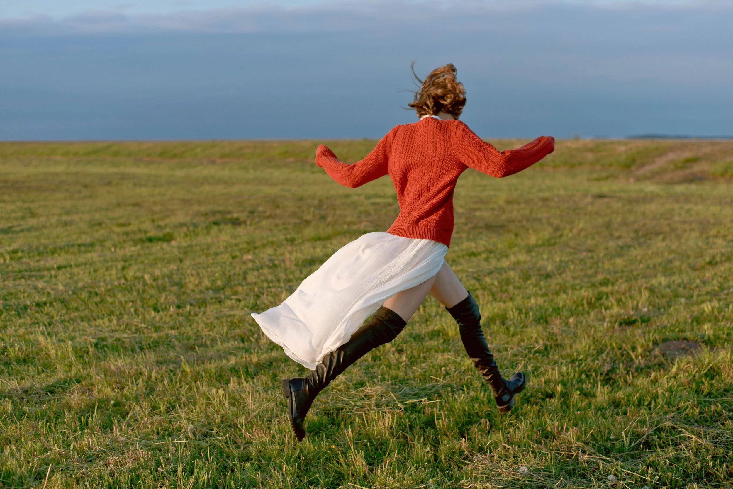 Girl in orange sweater and white skirt running through a green field