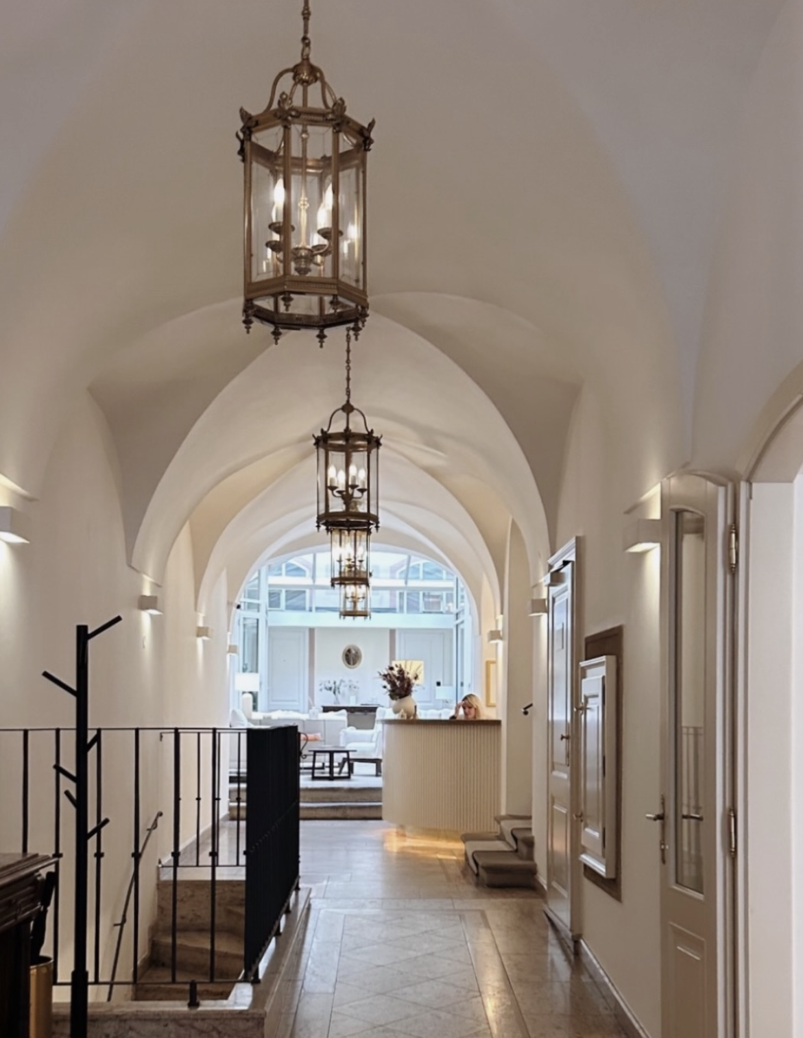 Elegant hallway with arched white ceiling, hanging lantern-style lights, and a view into a bright living room with large windows, furniture, and a person at a reception desk.