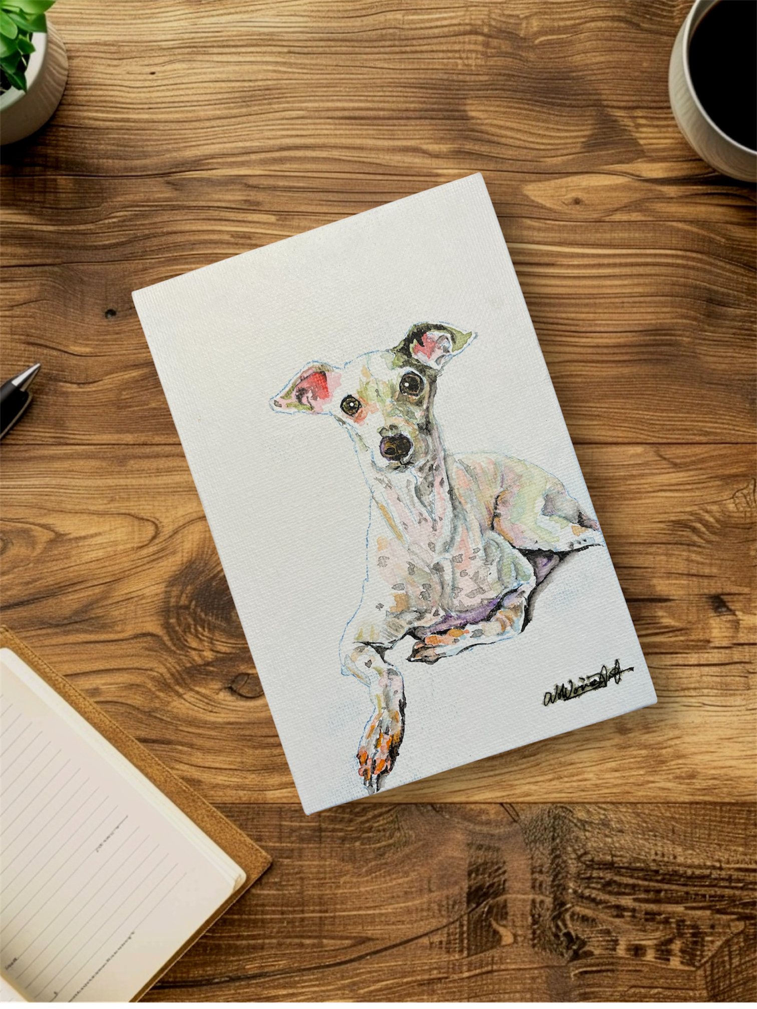 木の机の上に、犬の絵が描かれた白いキャンバスとノートとペン、コーヒーの入ったカップがあります。