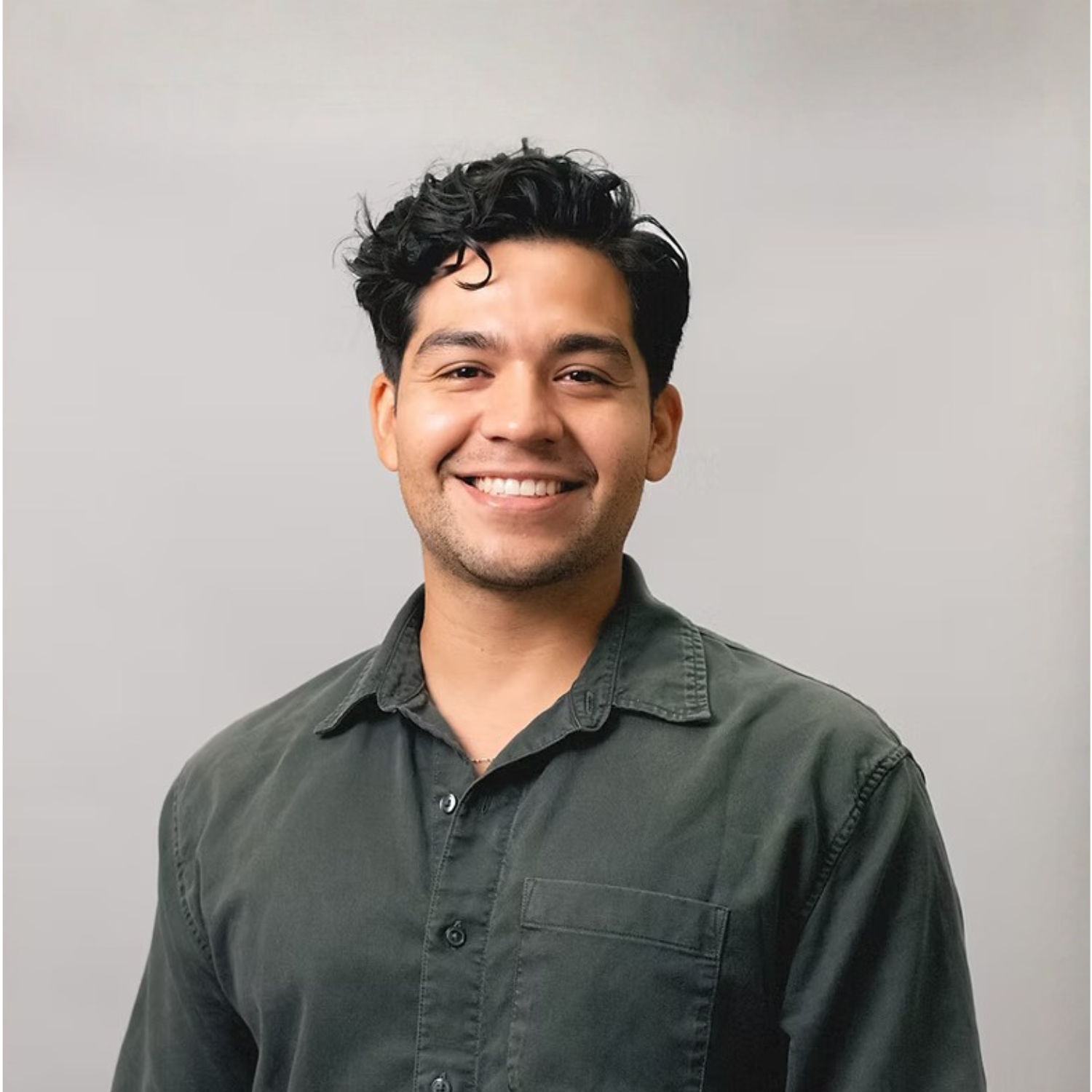 Smiling young man with dark curly hair wearing a dark button-up shirt against a light gray background.