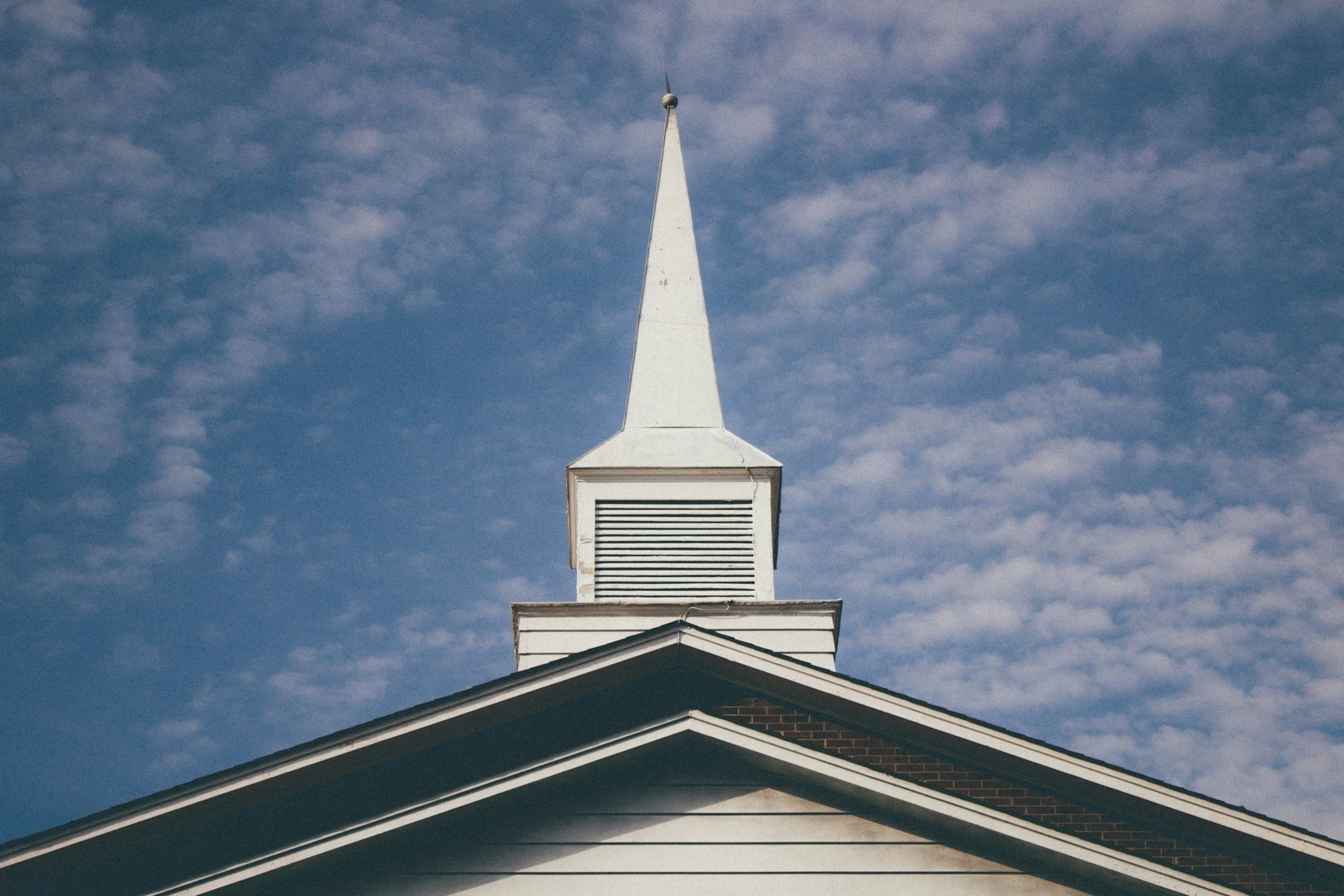 The steeple of a church building with a white spire against a partly cloudy blue sky.