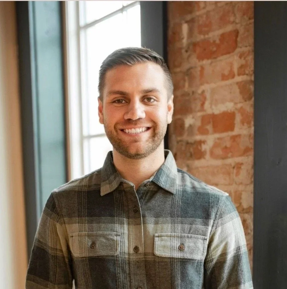 A smiling man with short brown hair and a beard, wearing a plaid shirt, standing indoors near a window with brick wall in background.
