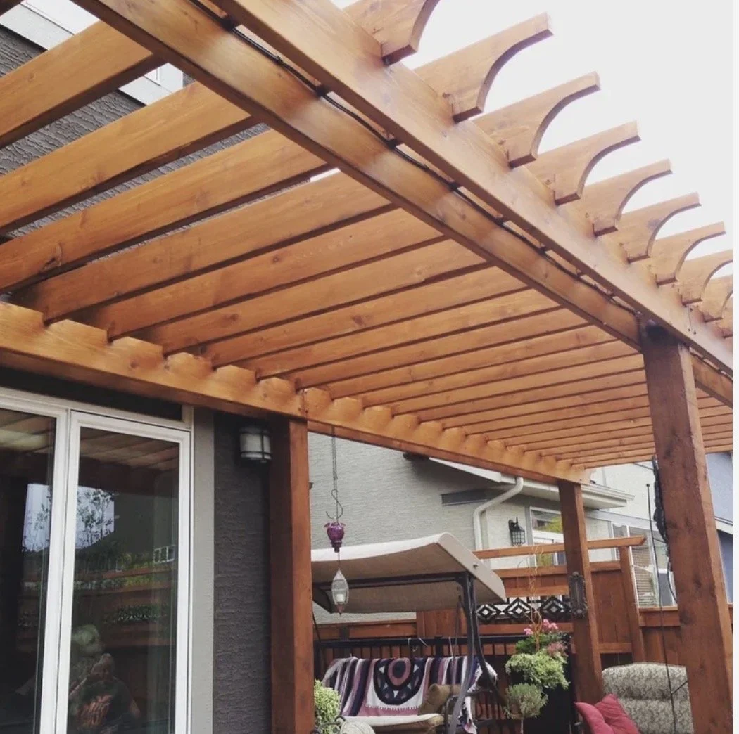 Wooden backyard patio pergola with slats and decorative curved ends, attached to house with sliding glass doors, outdoor furniture including a swing, potted plants, and string lights.