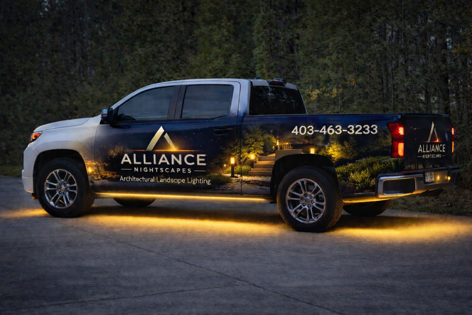 A pickup truck with a nighttime landscape lighting advertisement on its side, parked on a driveway with trees in the background. The truck displays the company name 'ALLIANCE NIGHTSCAPES' and a phone number, with lighting decoration in the design.