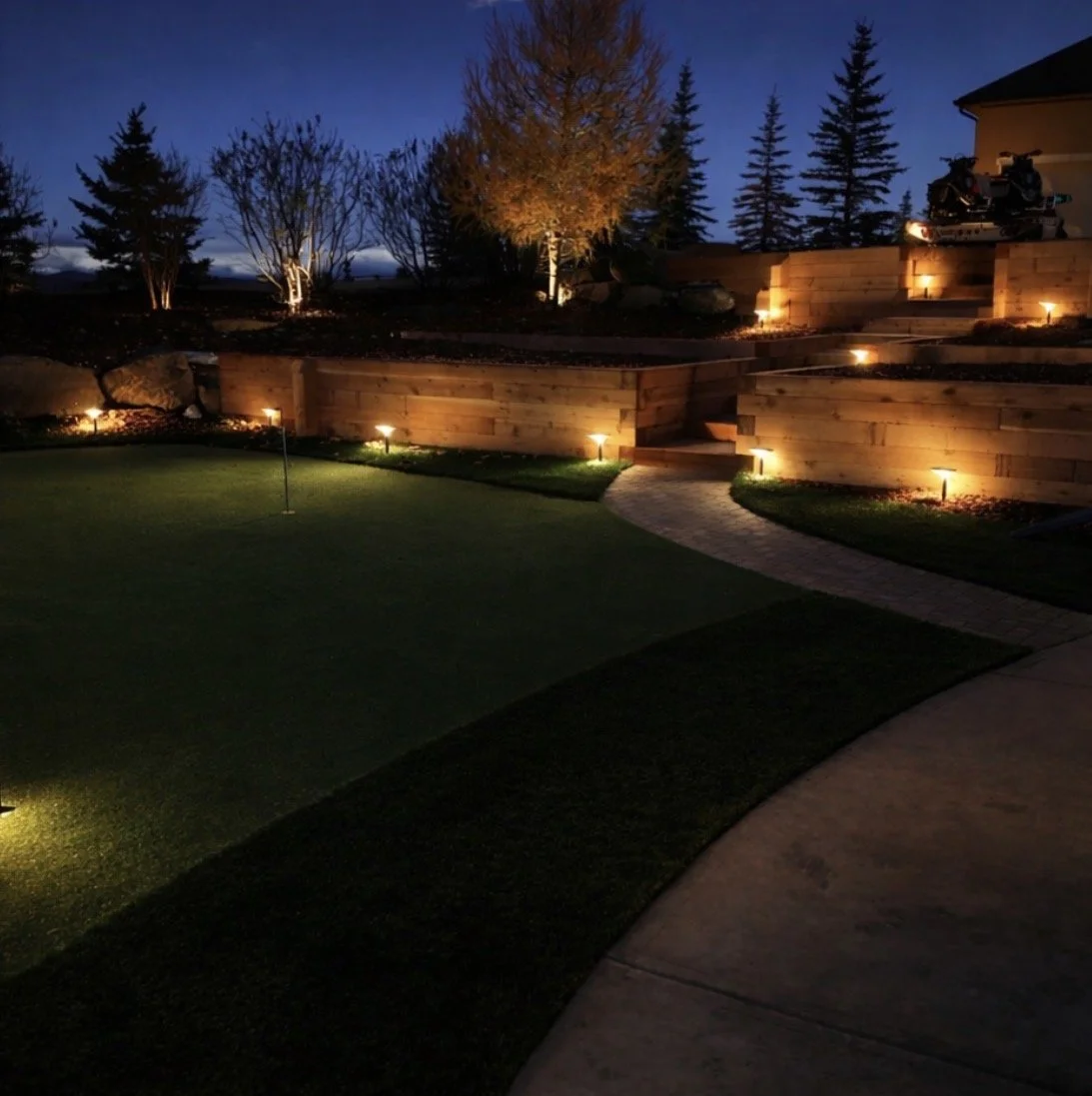 Nighttime view of a landscaped backyard with a green putting area, illuminated by ground lights, a curved paved pathway, and wooden retaining walls with planted trees and shrubs.