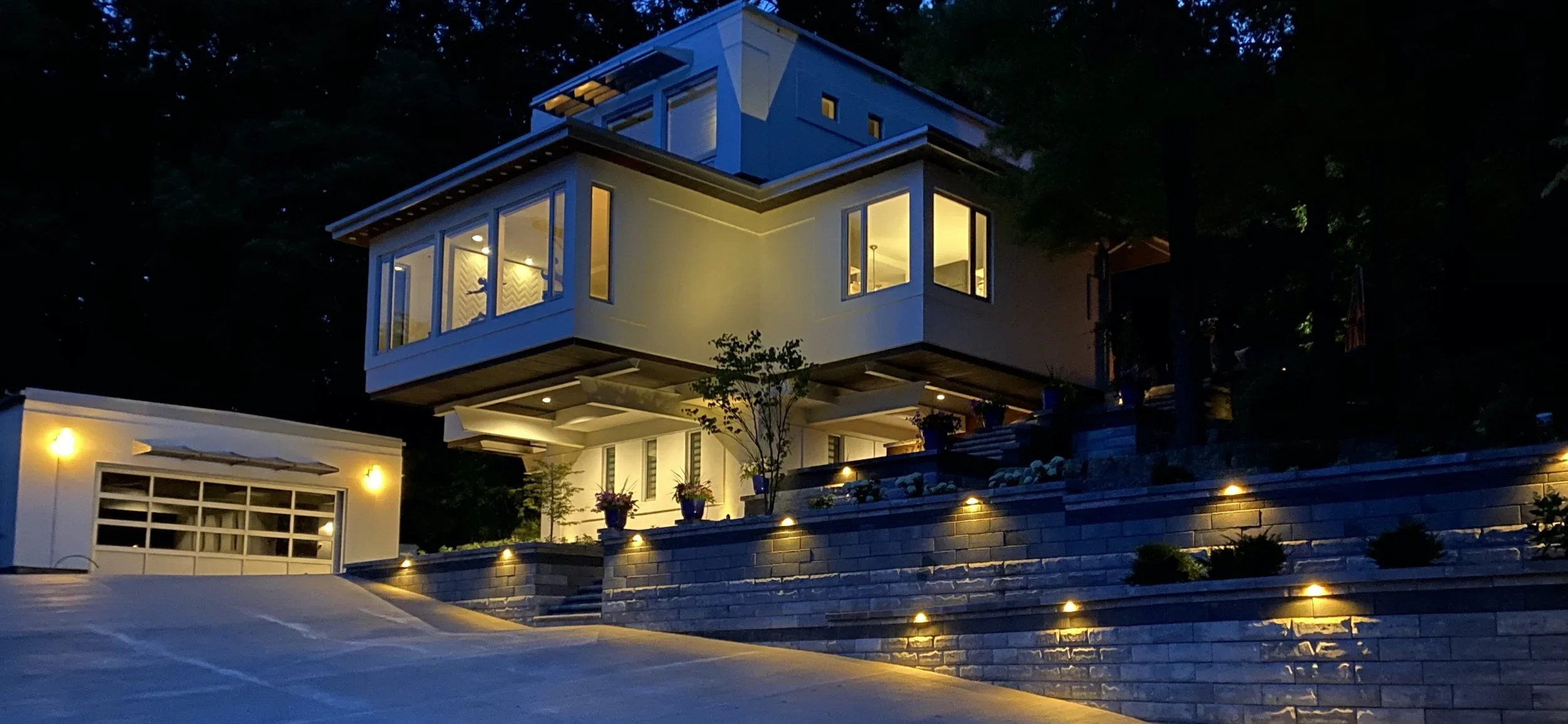 A modern house illuminated at night on a hillside with a multi-level design, large windows, outdoor lighting, and landscaping including potted plants and small bushes.