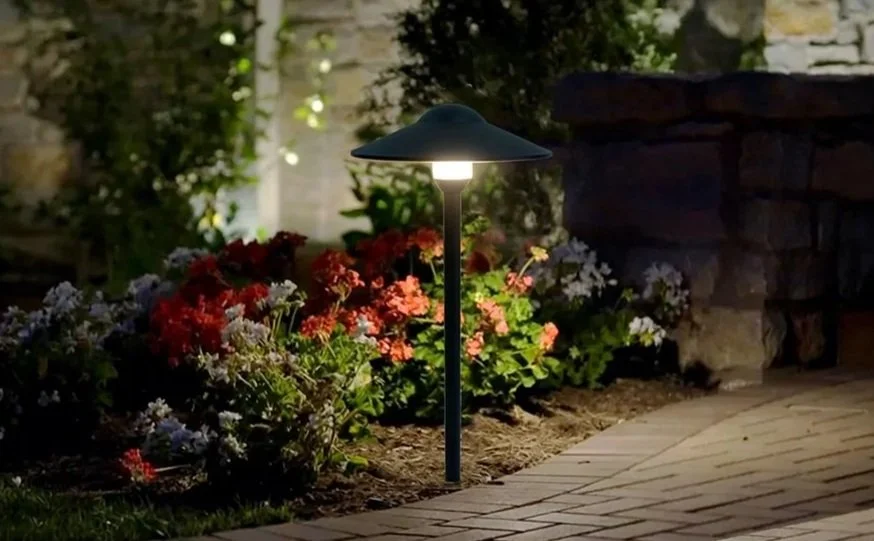 Outdoor garden pathway illuminated by a solar-powered lamp, surrounded by colorful flowers and stone wall.
