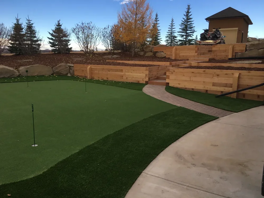 Backyard with a putting green, brick pathway, and wooden retaining walls, trees, and a small building with a tracked vehicle on the upper level.
