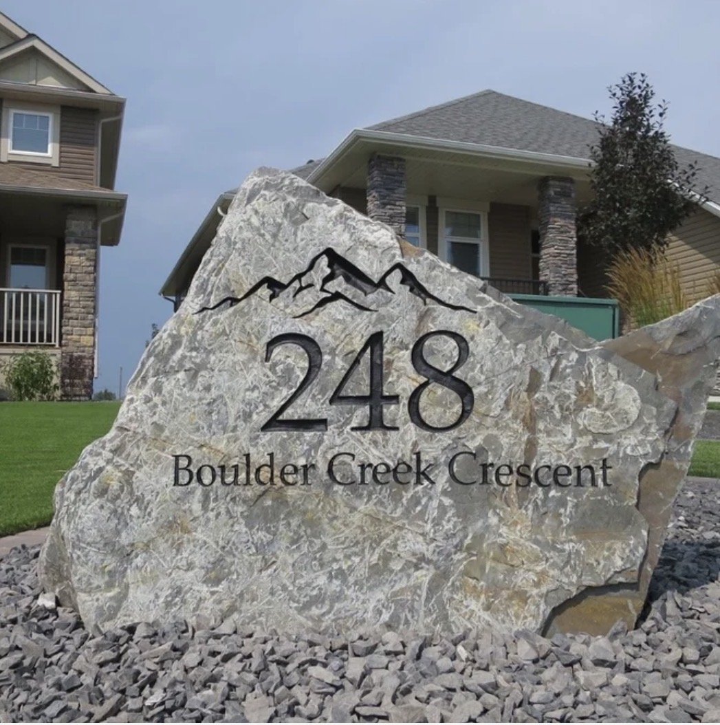A large decorative stone with the house number '248' and the street name 'Boulder Creek Crescent' engraved on it, situated in the front yard of a house with a lawn and neighboring homes visible in the background.