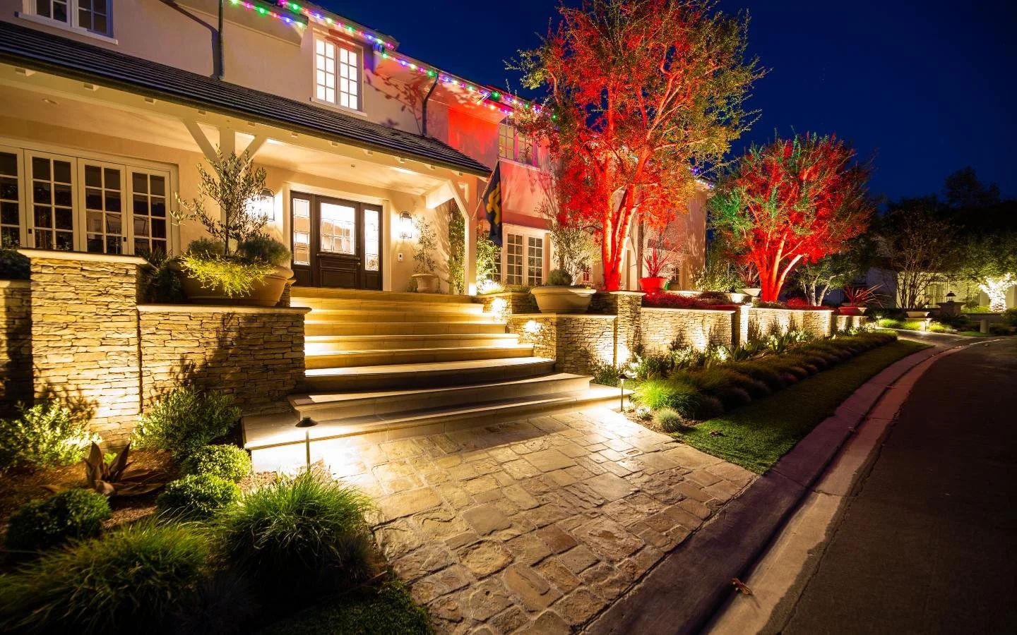 A well-lit house with steps leading up to the front door, decorated with colorful string lights and illuminated trees during nighttime.