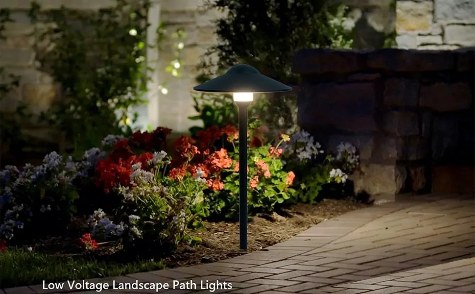 Outdoor landscape scene at night with illuminated low voltage pathway lights along a brick walkway, surrounded by colorful flowers and stone wall.