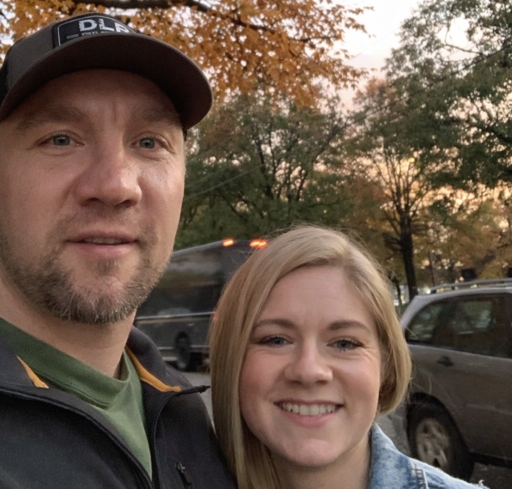 A man and woman taking a selfie outdoors during sunset, with trees and cars in the background.