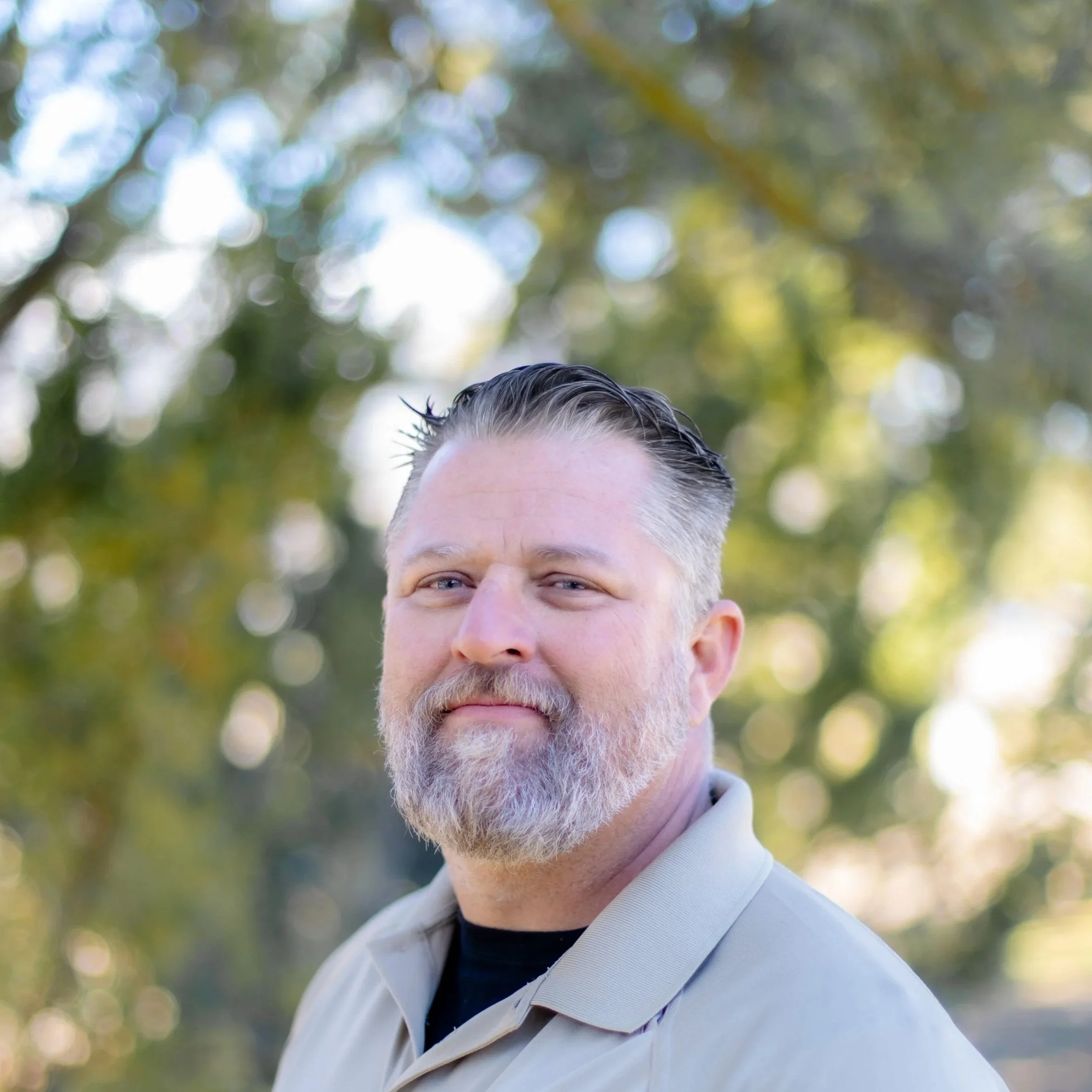 A middle-aged man with gray hair and a beard, wearing a light-colored shirt, standing outdoors with blurred green and yellow foliage in the background.